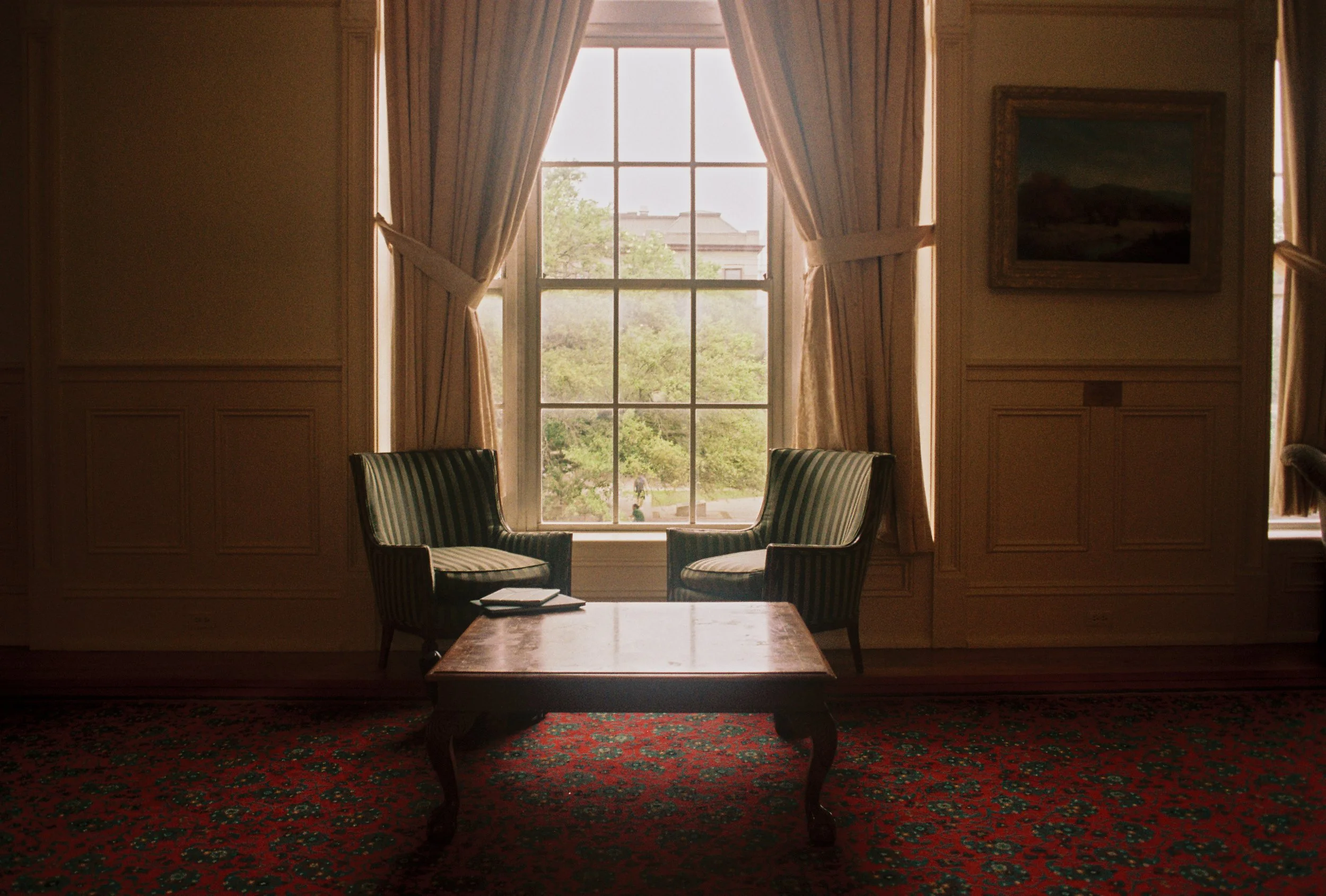 A cozy sitting room with two green and white striped armchairs facing each other, a wooden coffee table between them, and a large window with cream-colored curtains revealing a view of trees and a building outside.