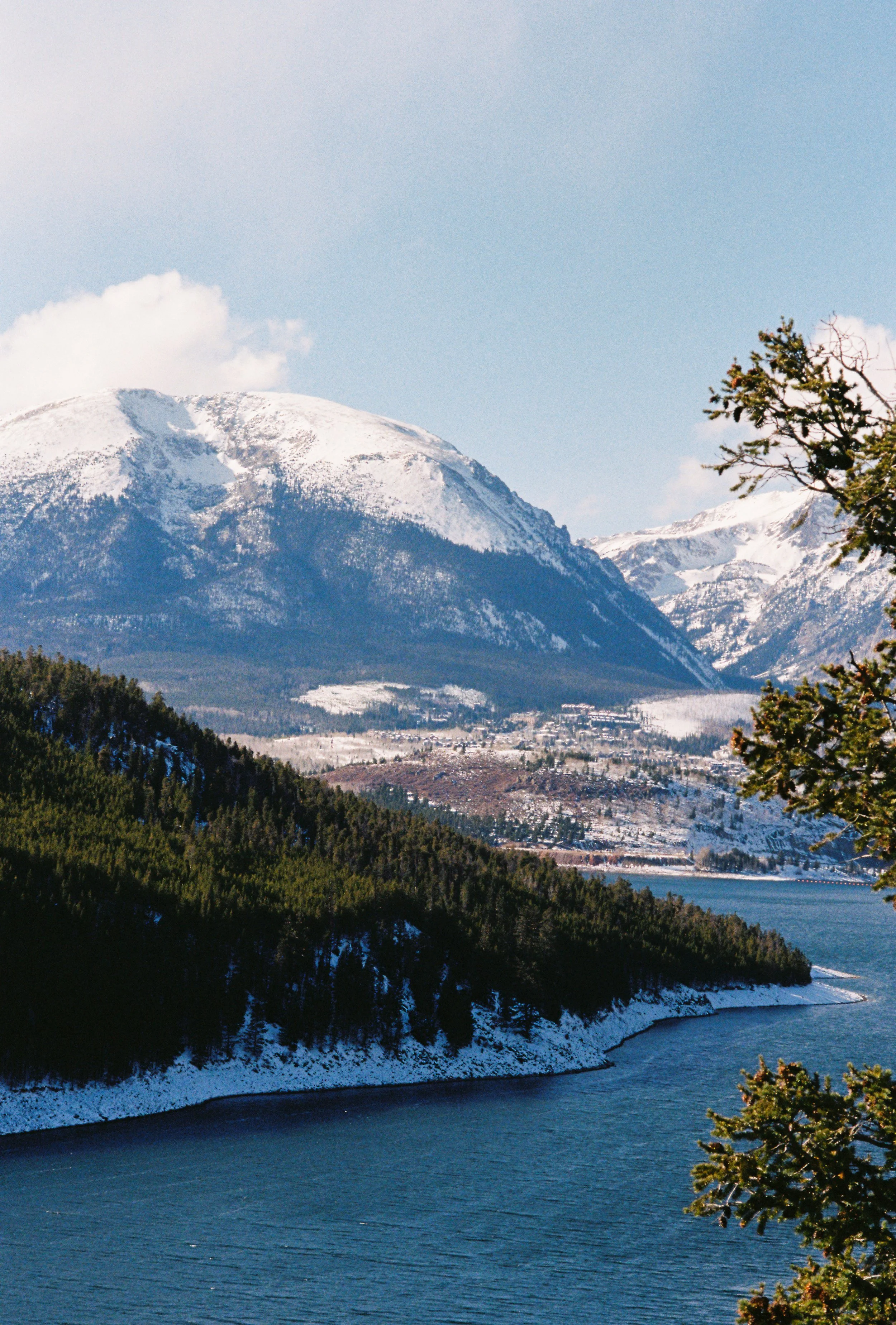 Snow-covered mountains surrounding a body of water with green pine trees on the shoreline and some on the right side of the image.