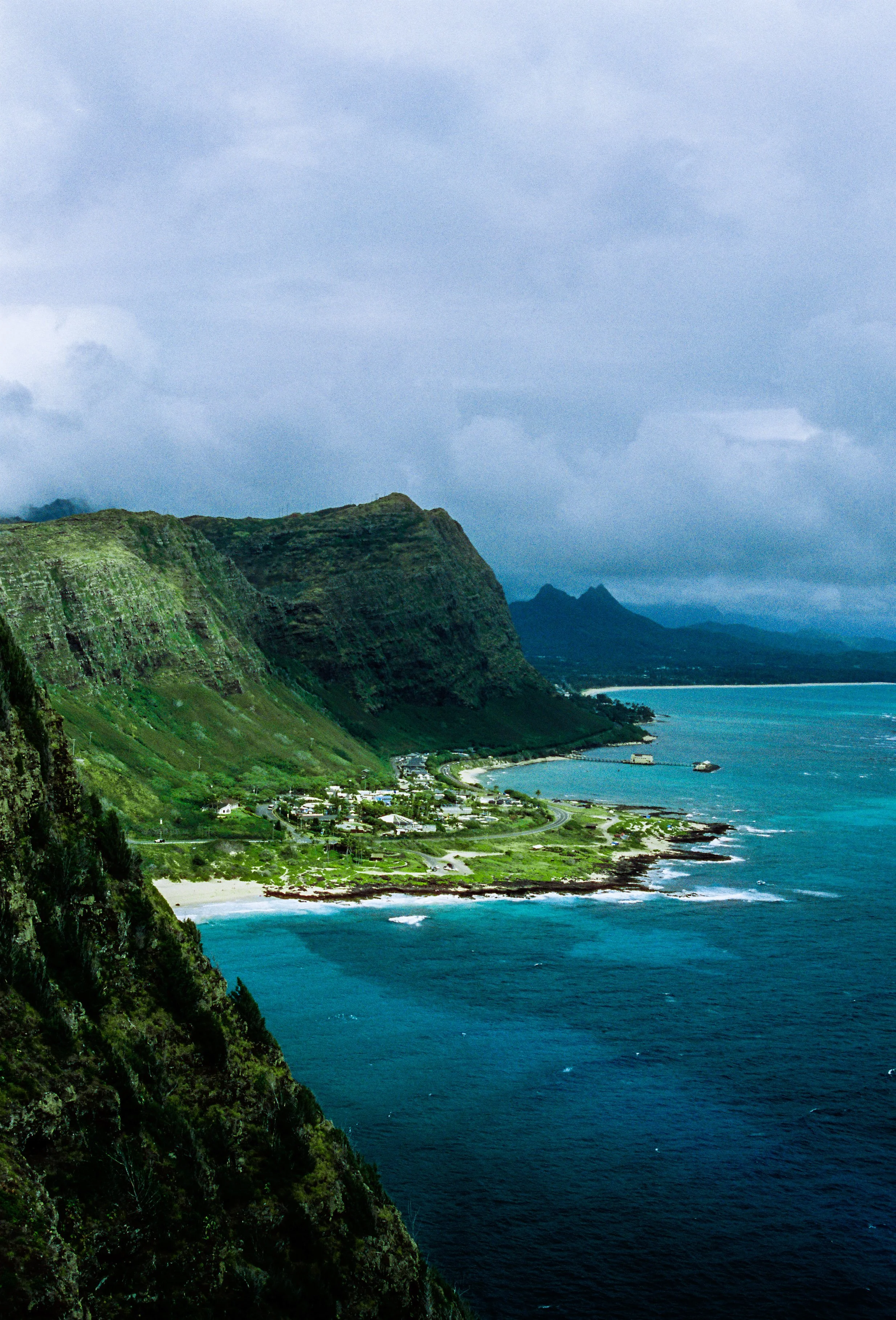 Scenic view of green mountains and coastline with a small town and ocean in the distance under cloudy sky.