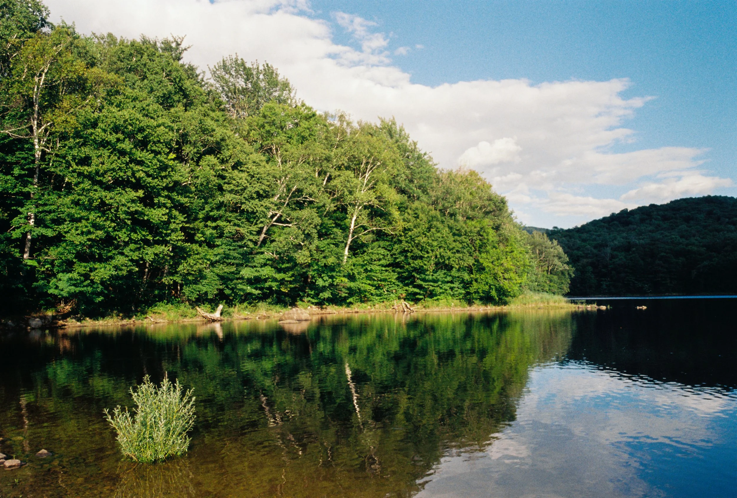 A river with calm, reflective water surrounded by lush green trees on the left and a hill covered with greenery in the background on the right, under a partly cloudy sky.