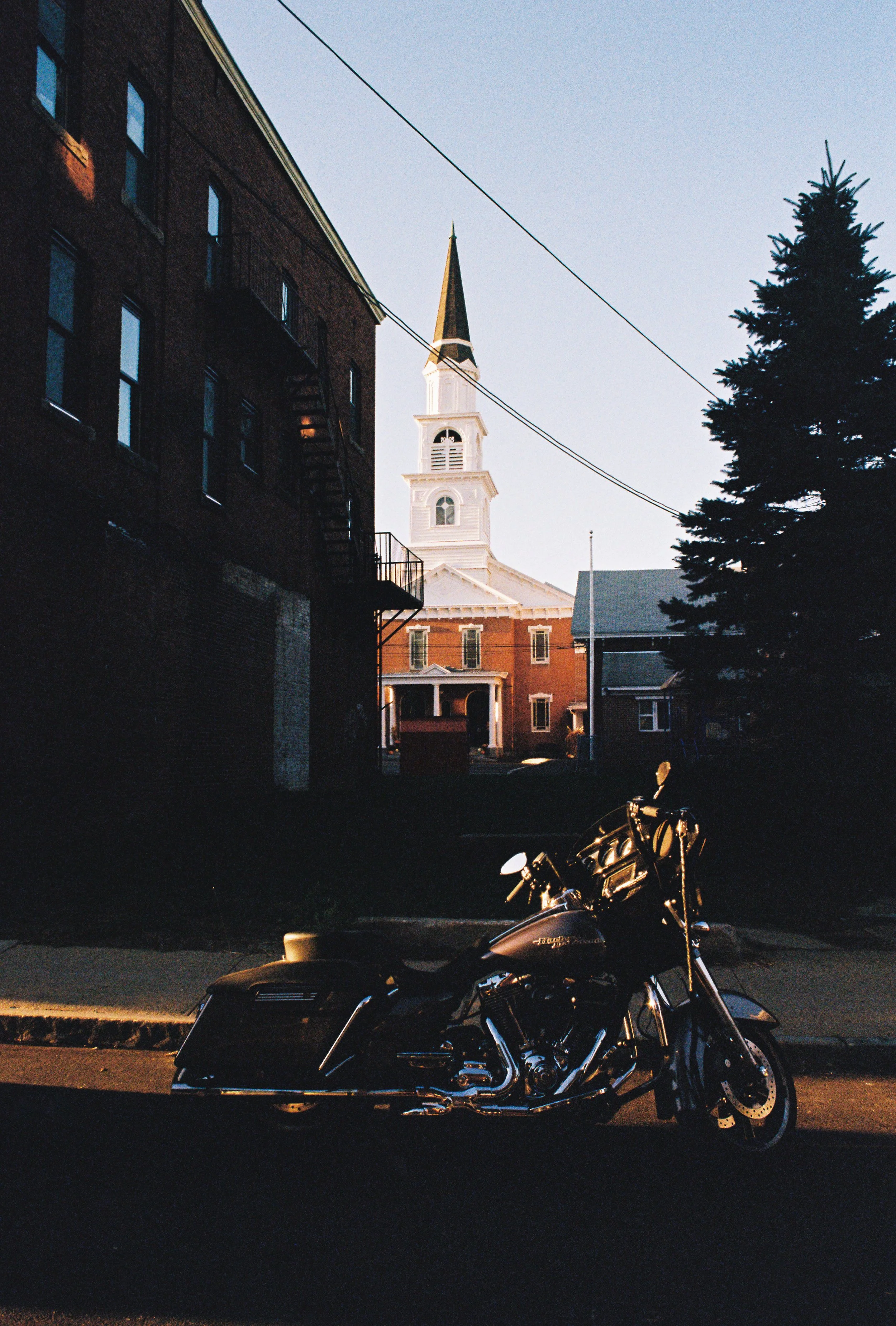A motorcycle parked on the street with a church and trees in the background, illuminated by late afternoon or early evening light.