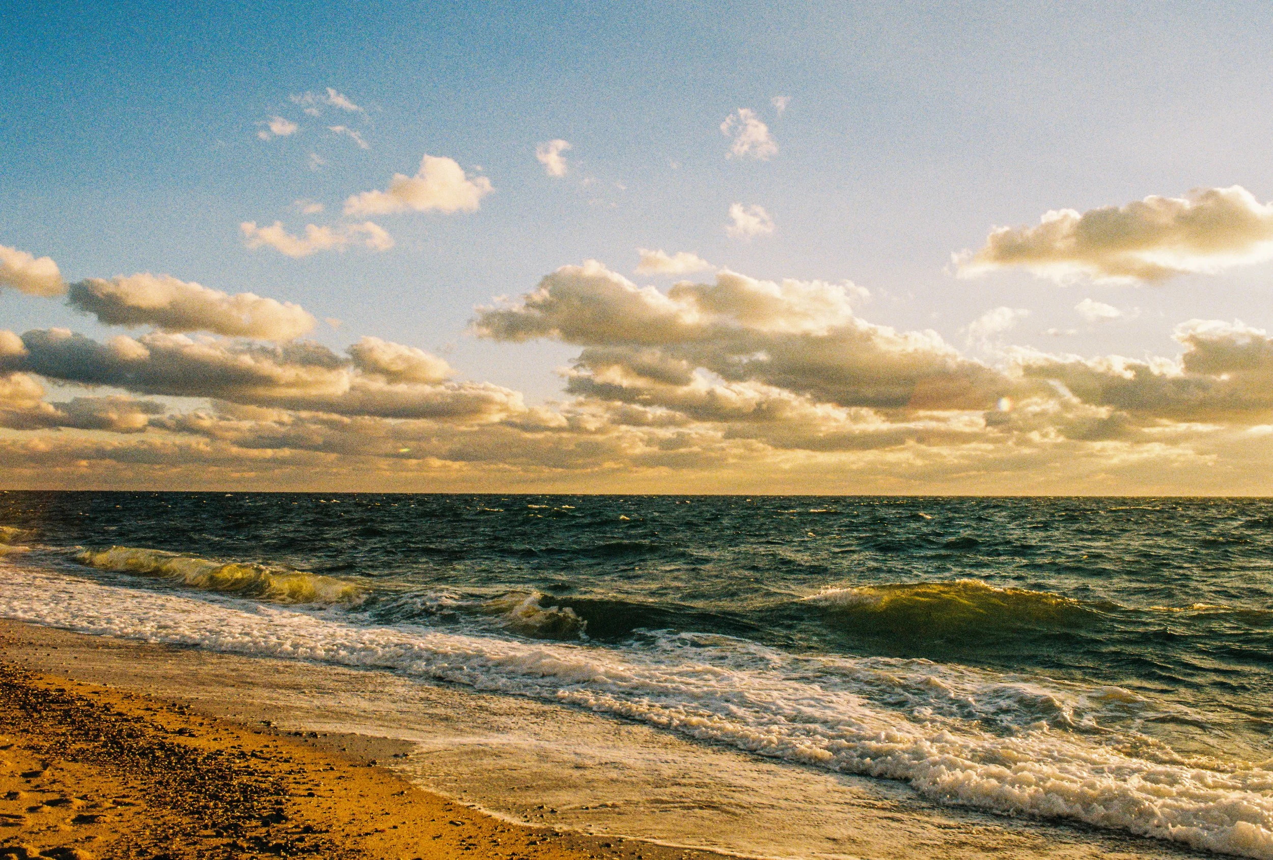 A view of the ocean at sunset or sunrise with waves crashing on the sandy beach, and a sky filled with fluffy clouds.