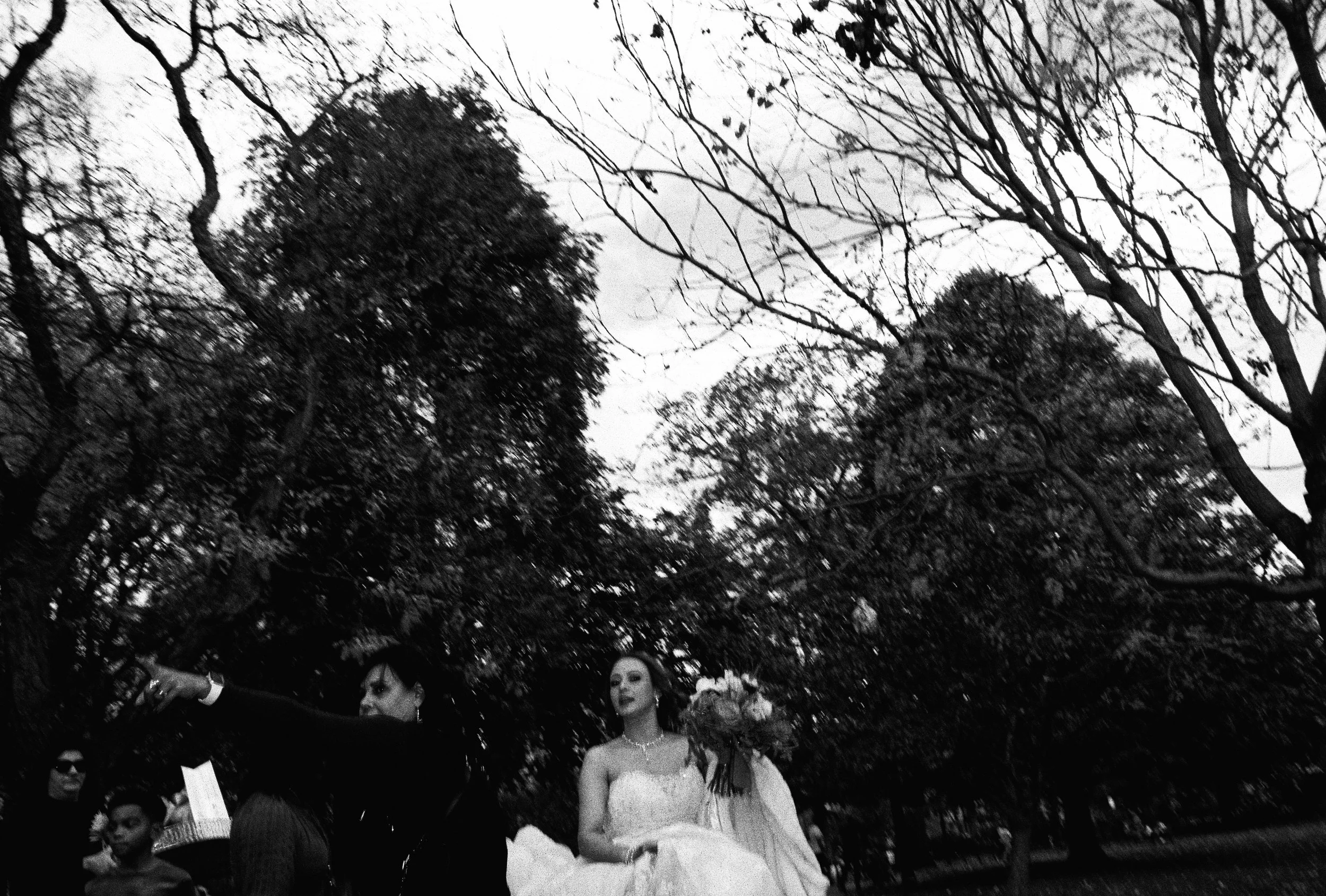 Black and white photo of women and children outdoors among leafless trees, one woman in a wedding gown holding a bouquet, others dressed formally.