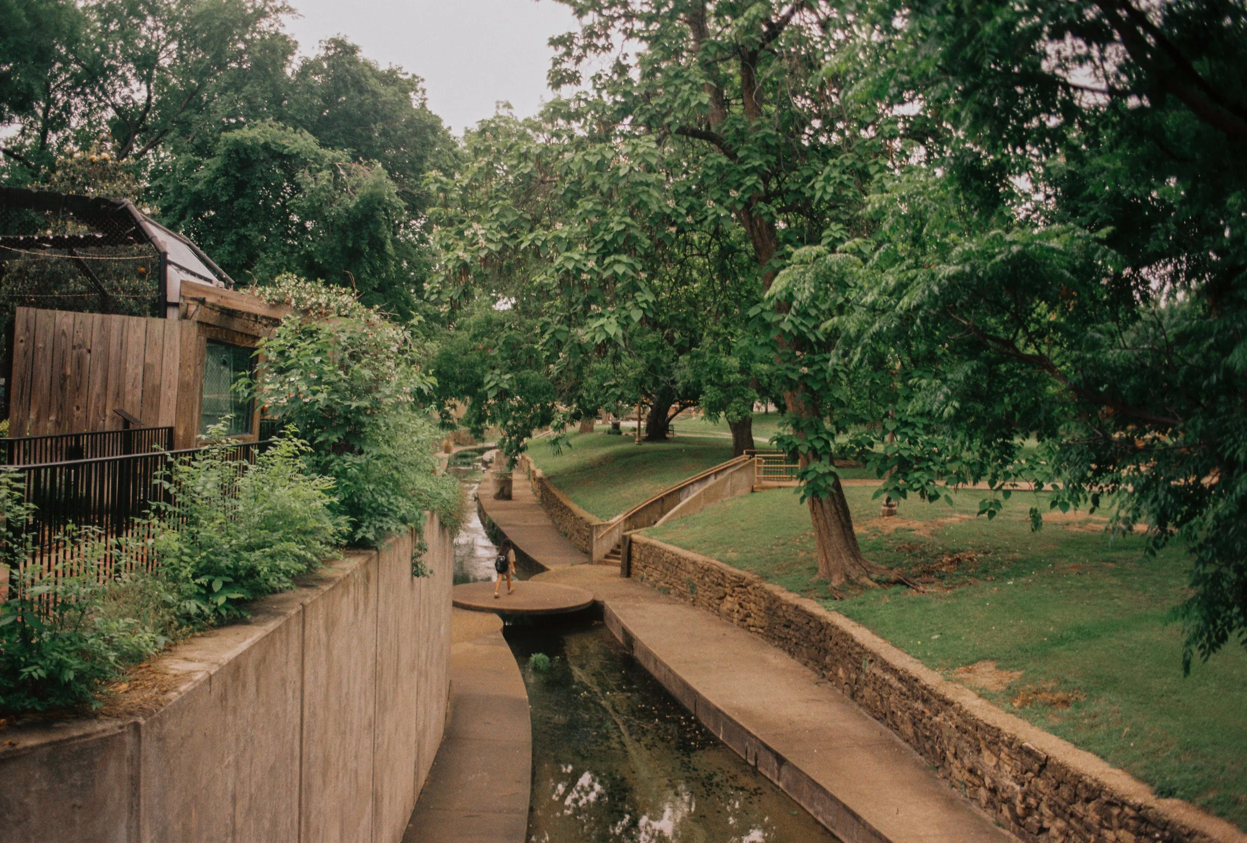 A scenic park scene with a narrow creek running through it, surrounded by concrete and stone walls, lush green trees, and a person walking along the pathway.