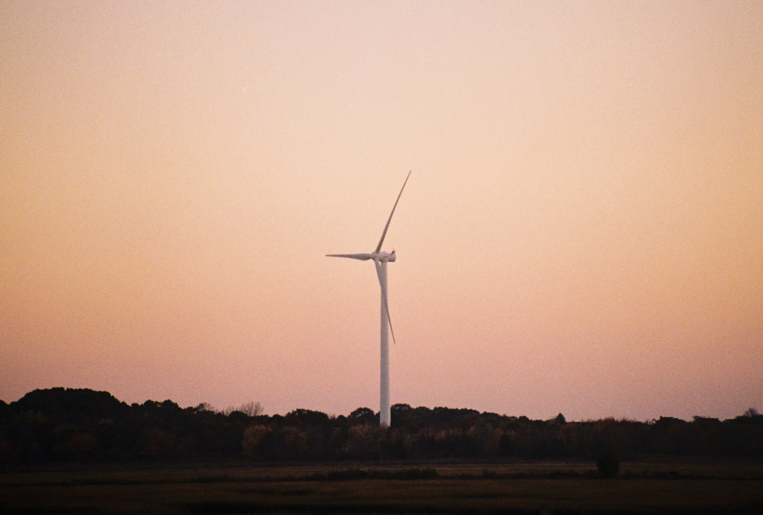 A landscape with a wind turbine against a pinkish-orange sky at dusk or dawn, with a line of trees in the foreground.