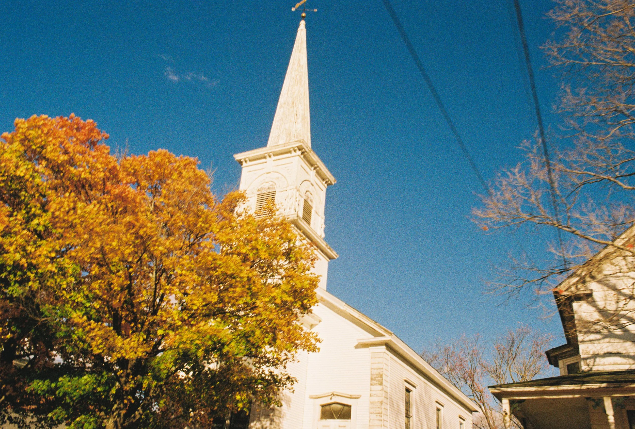 A white church with a tall, pointed steeple under a clear blue sky, surrounded by trees with autumn foliage.