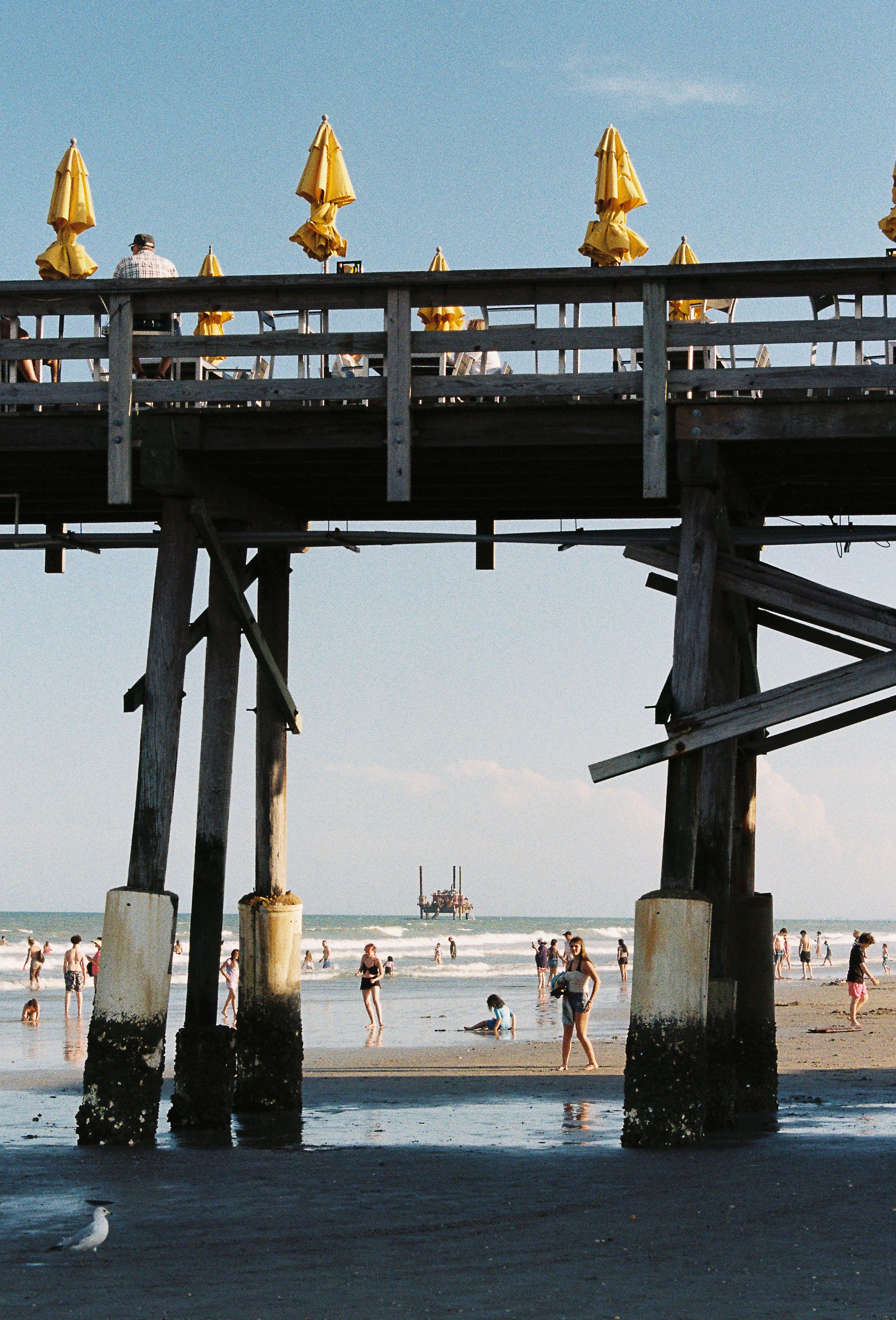 A beach scene viewed through a large wooden pier with yellow umbrellas on the upper deck. People are walking and sitting on the sand and in the water. An offshore oil platform is visible in the distance against a clear blue sky.
