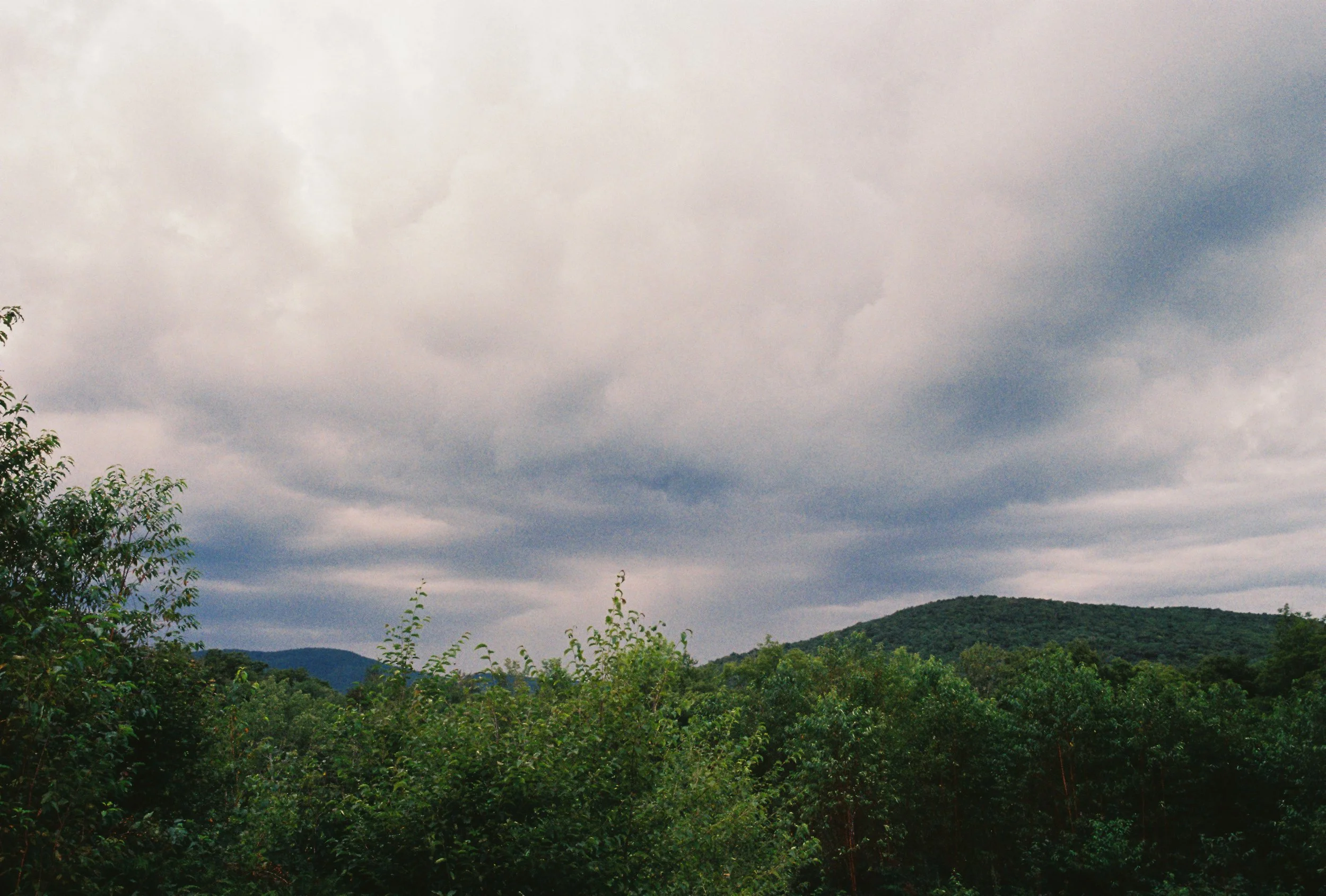 Overcast sky with dark clouds over a green wooded hillside.