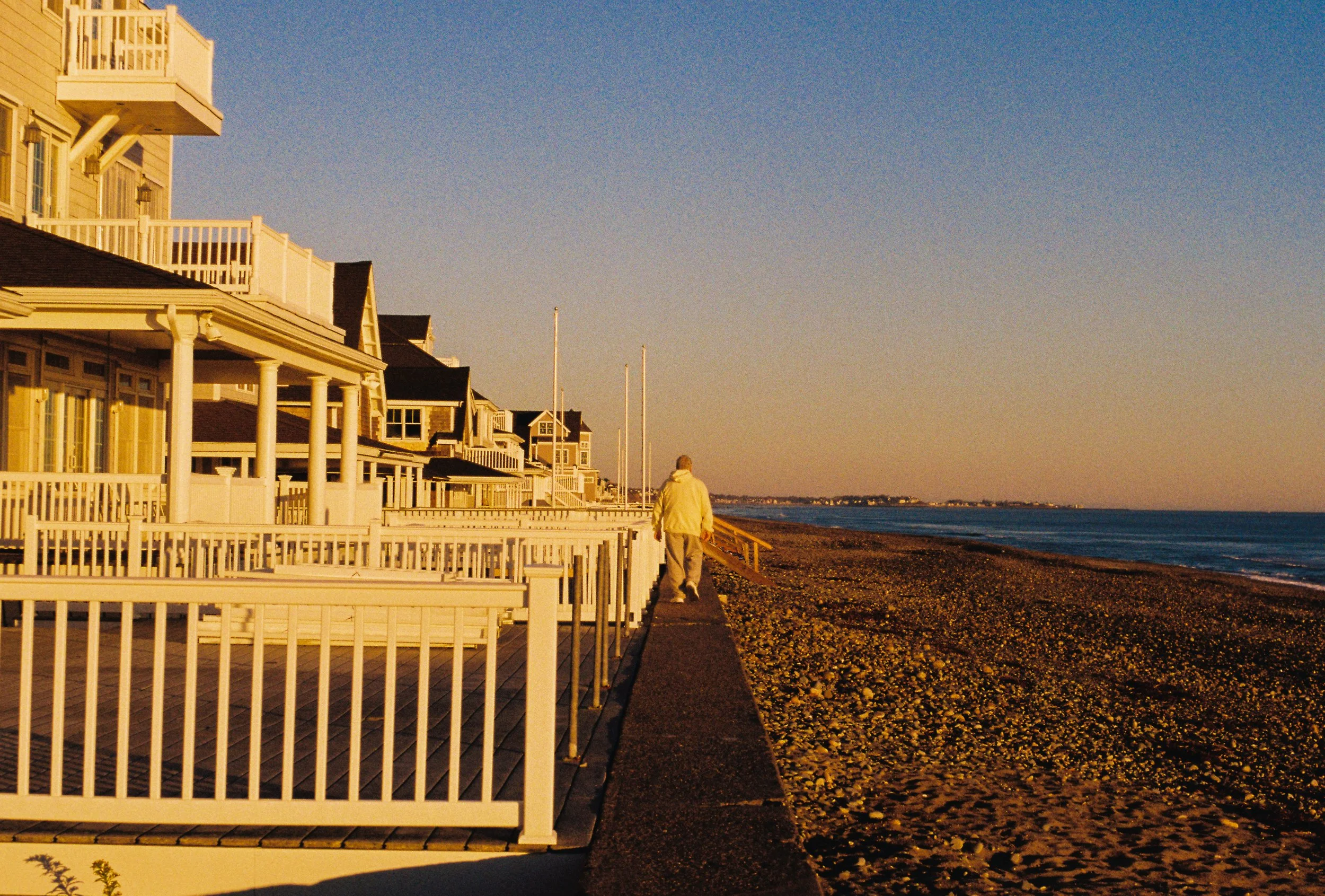 A person in a yellow jacket walking along a seaside walkway at sunset, with beach houses on the left and the ocean on the right.