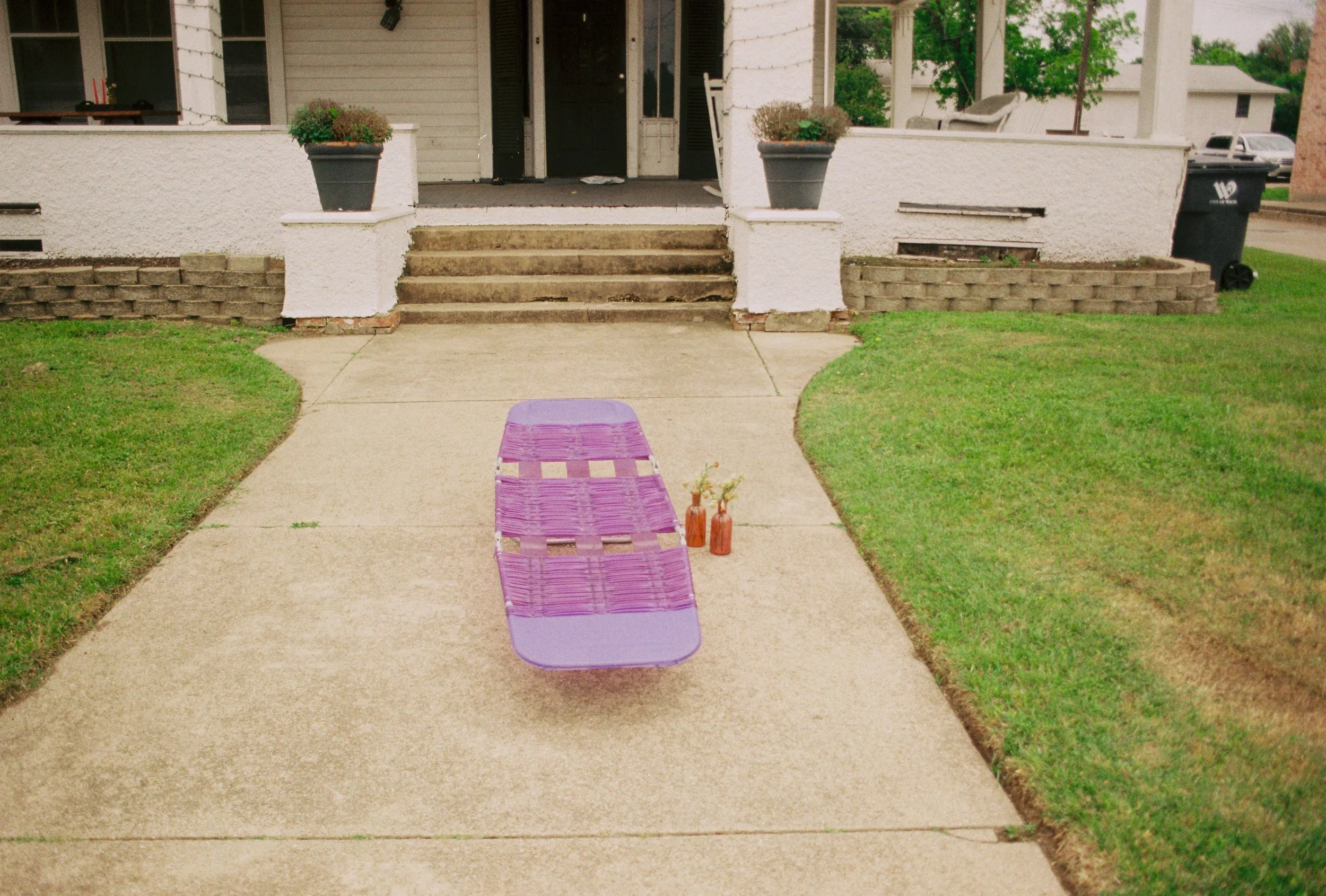 Purple lounge chair on a concrete sidewalk in front of a house with steps, potted plants, and green lawn.