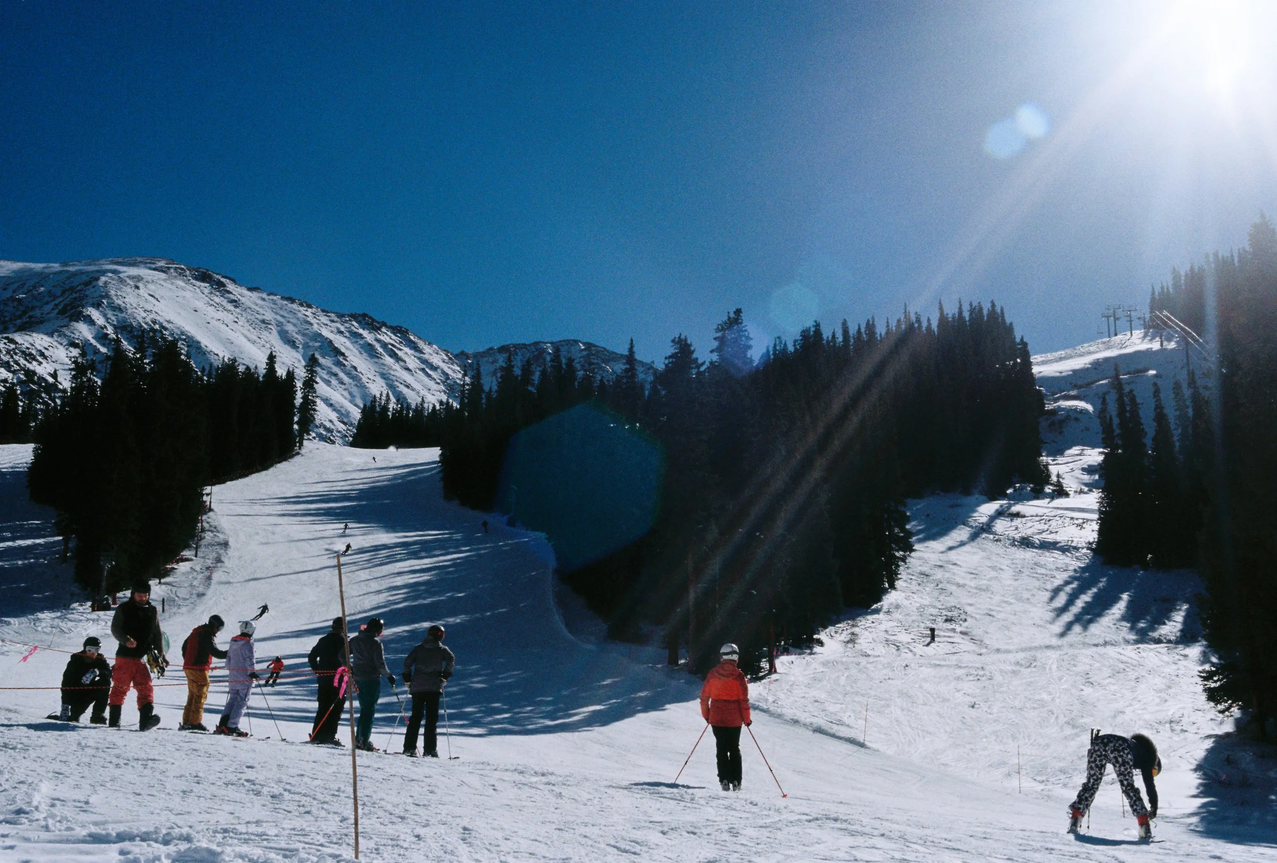 Group of people waiting in line to ski on a snowy mountain slope, with tall trees and mountain peaks in the background, under a bright sunny sky.
