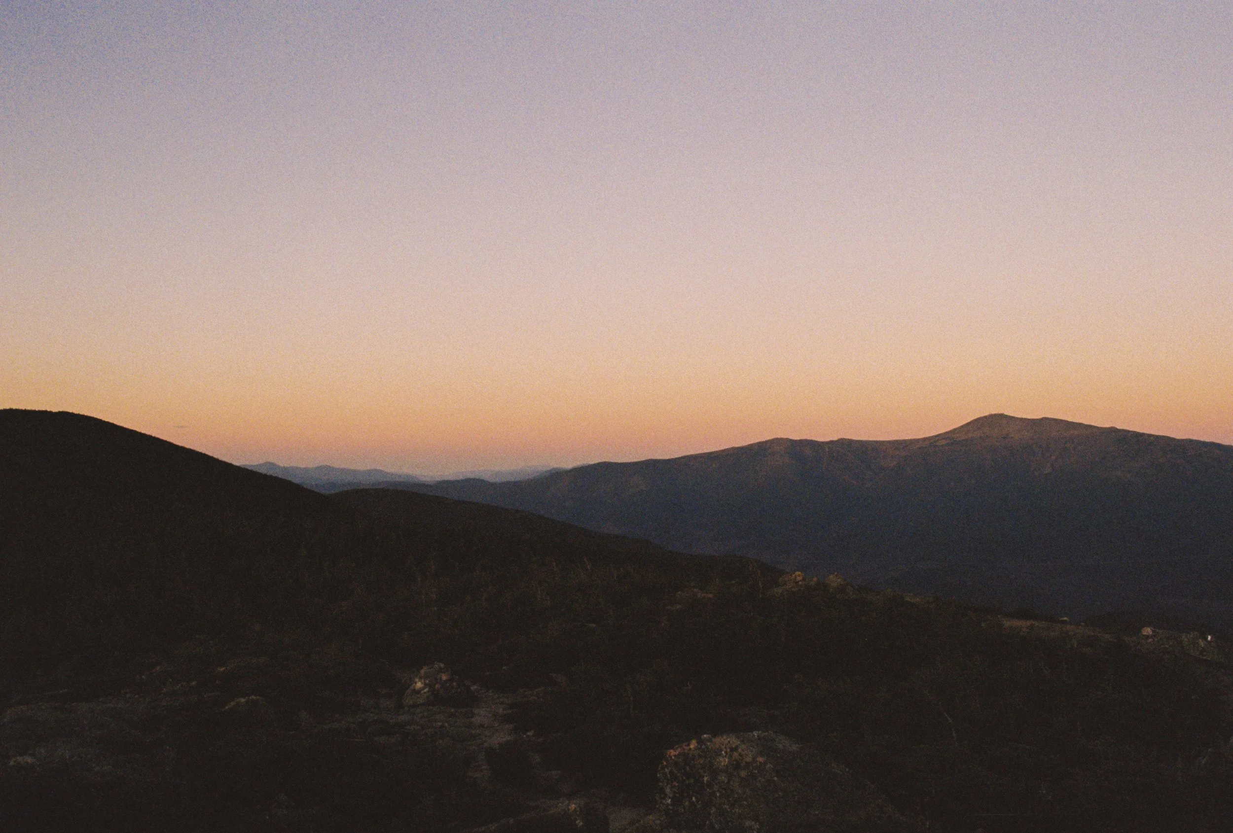 Mountain landscape at sunset with dark hills in the foreground and lighter, distant mountains under a mostly clear sky.