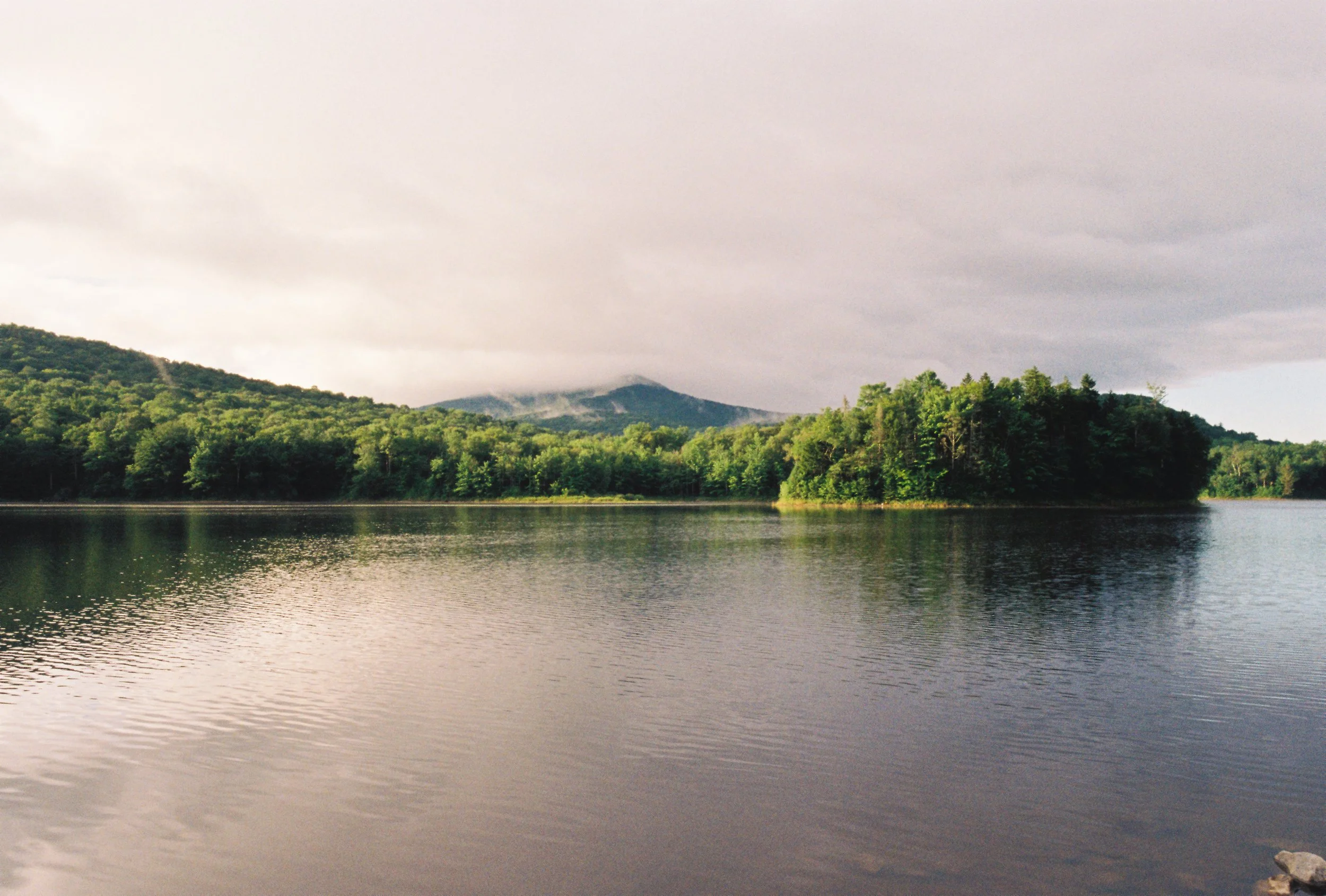Scenic view of a lake with calm water, surrounded by lush green trees and hills in the background under a cloudy sky.