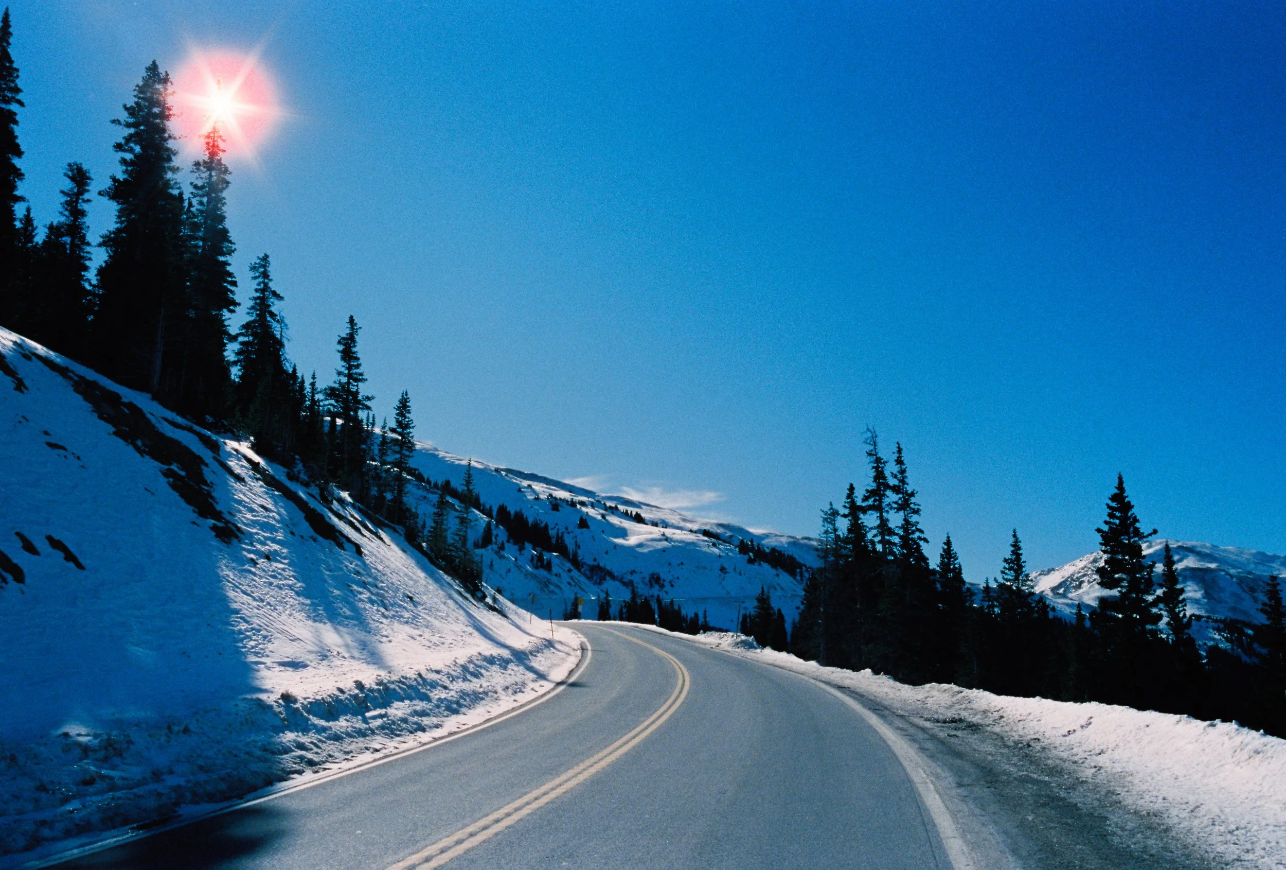 Snow-covered mountain road winding through a forested landscape under a clear blue sky, with the sun shining brightly.