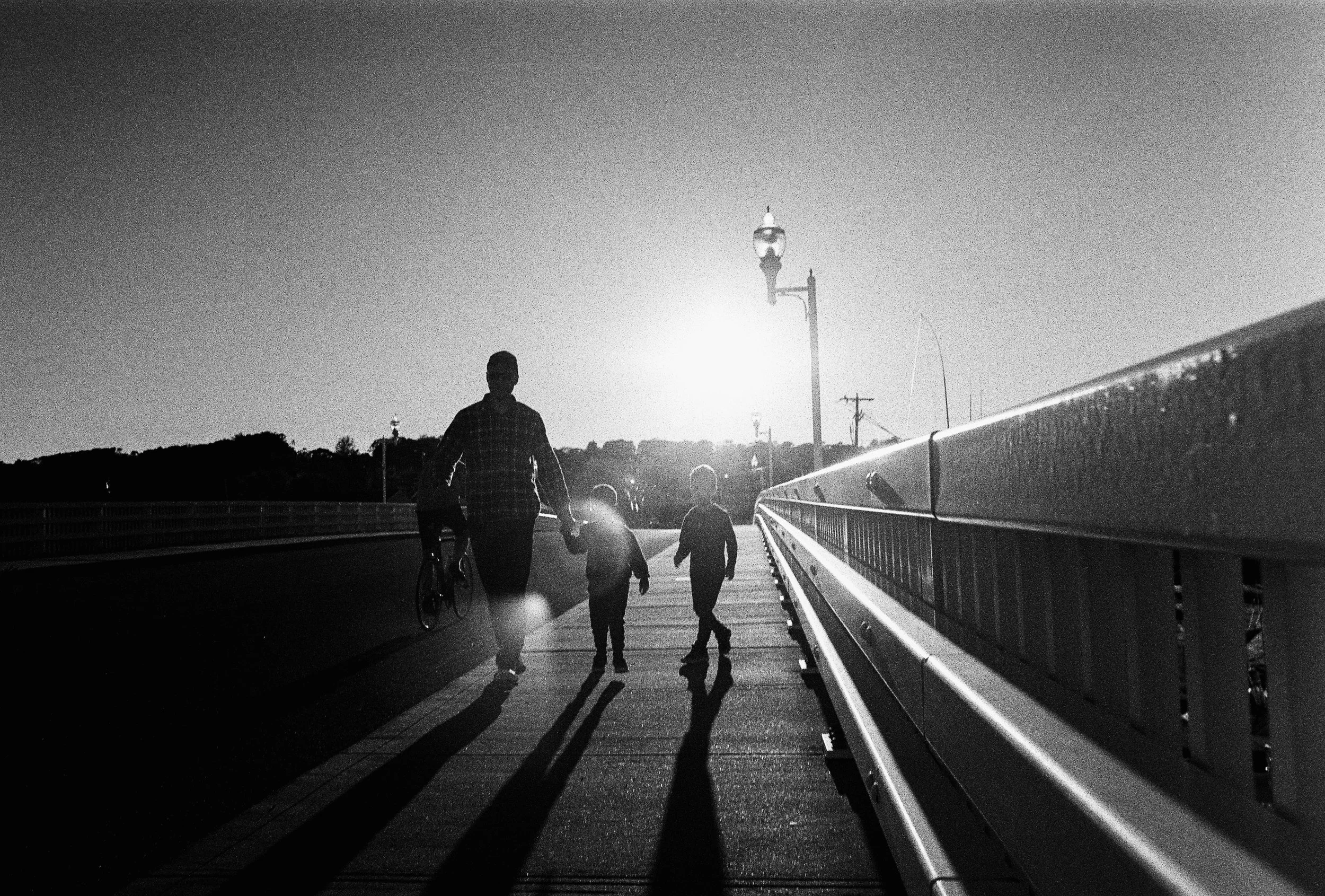 A silhouette of a person and two children walking on a bridge at sunset, with a streetlamp and a bicyclist in the background.