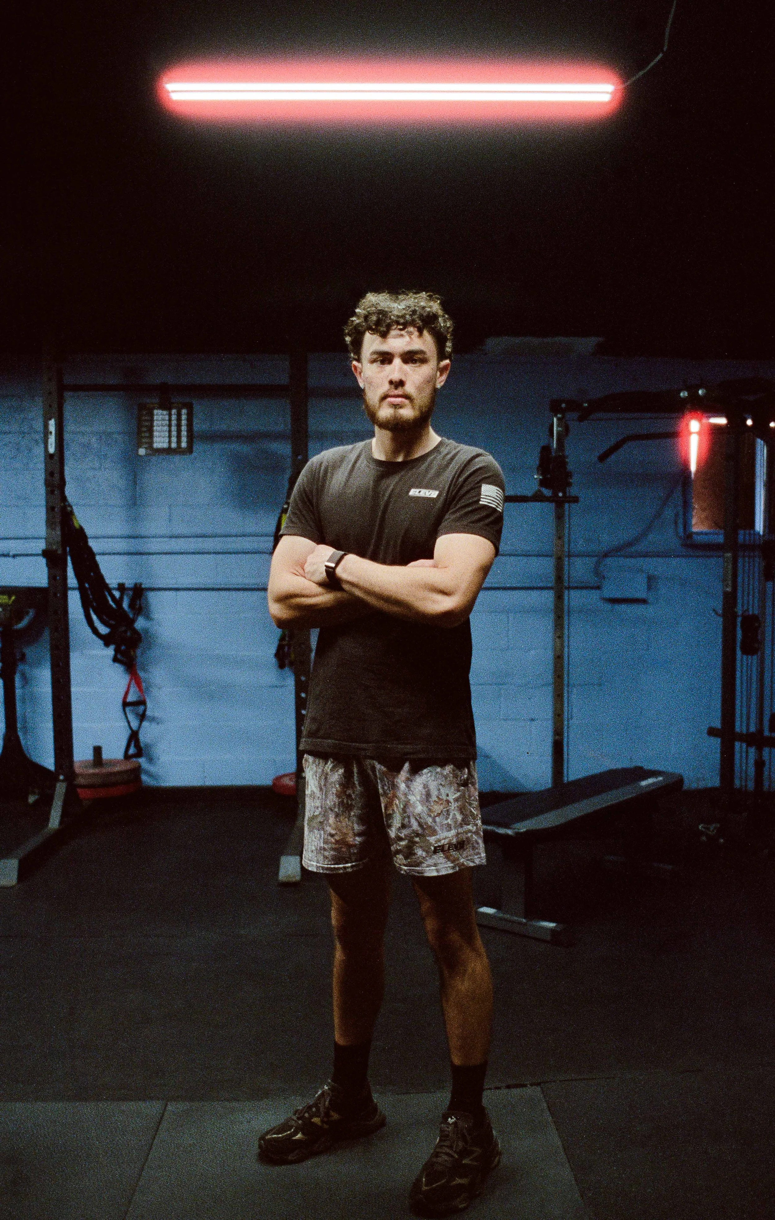A man with curly hair and a beard standing in a gym with arms crossed, wearing a black t-shirt, camouflage shorts, and running shoes.