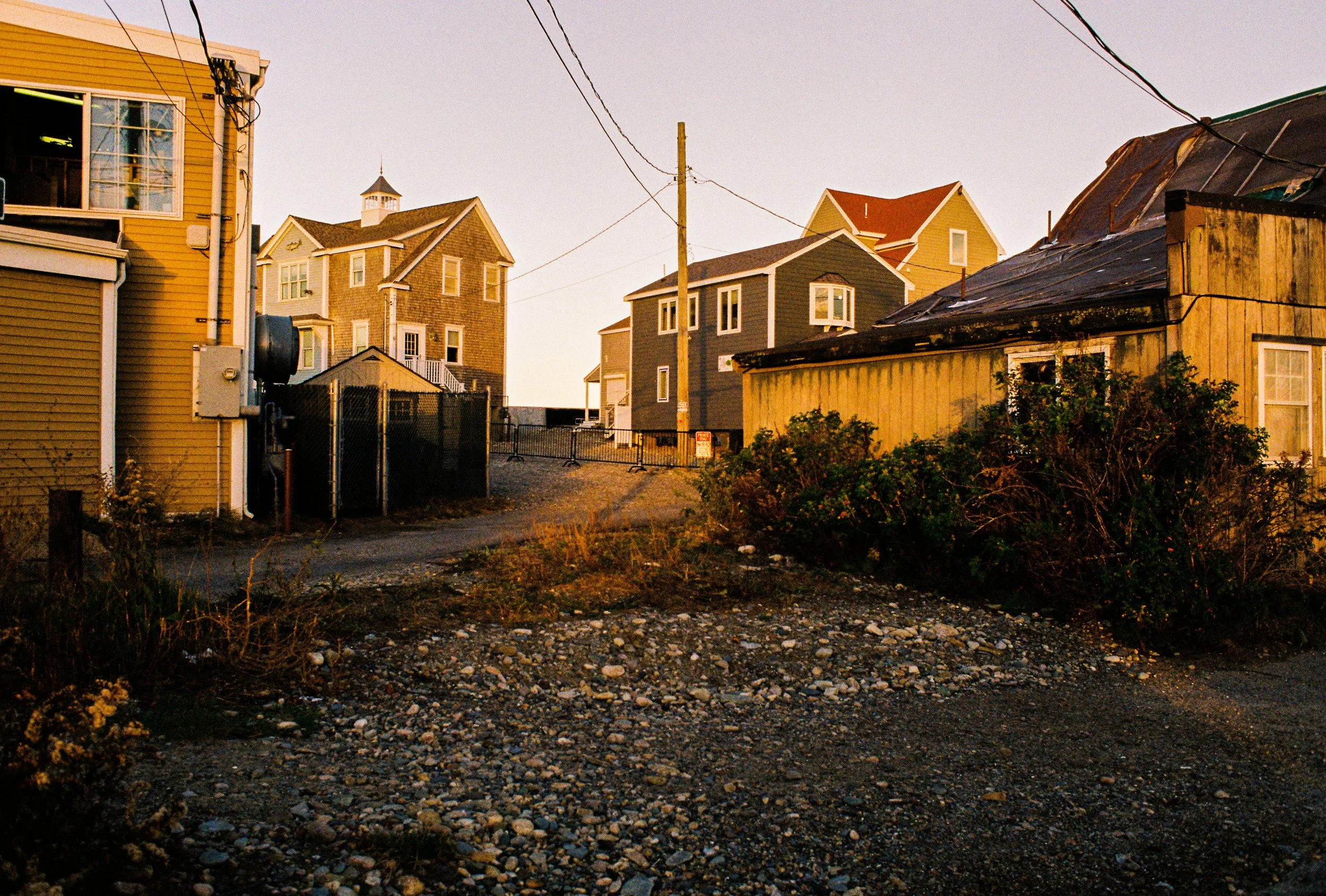 A dirt and gravel alleyway between colorful residential houses during sunset with overhead power lines.