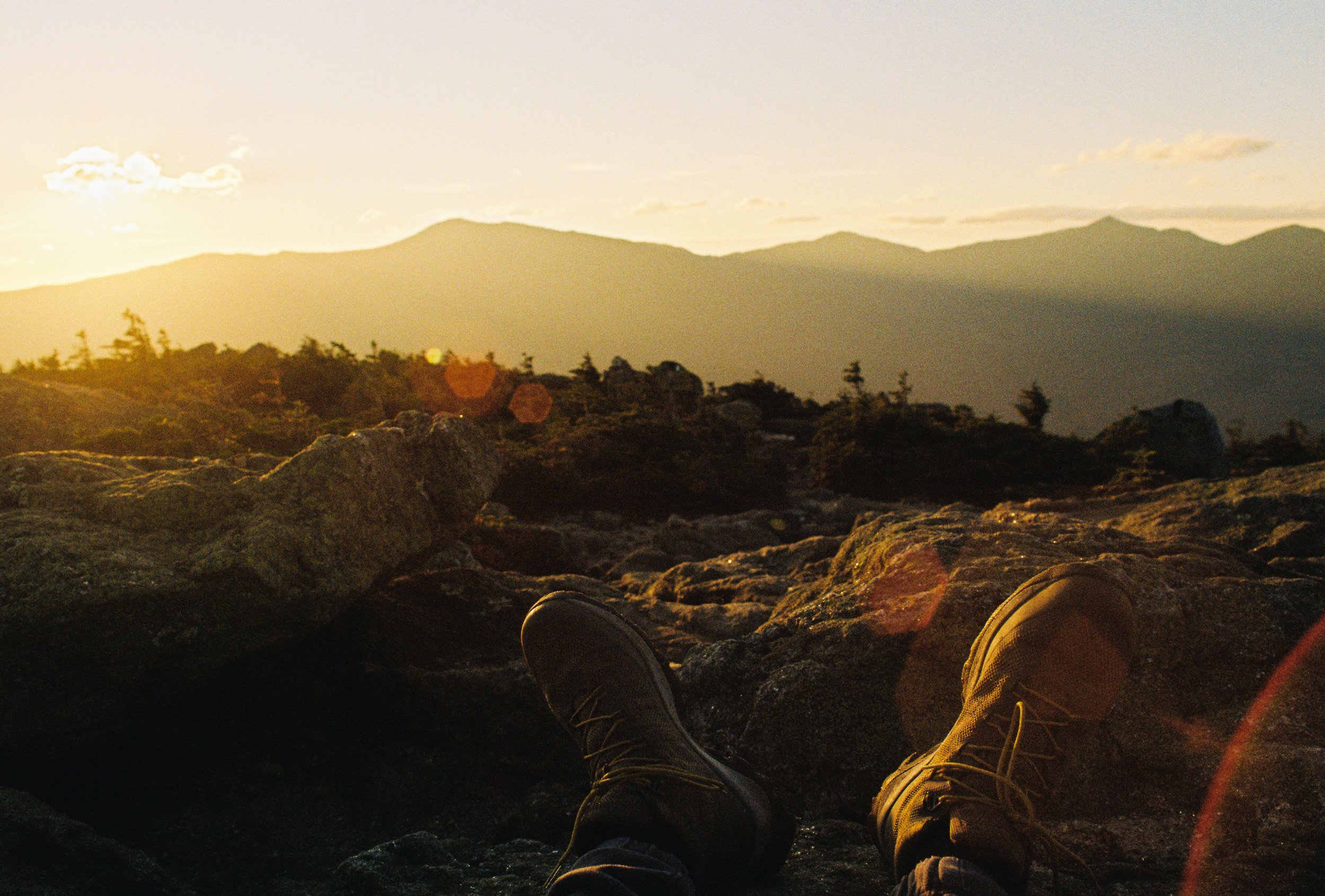 Person reclining on rocky terrain during sunset or sunrise with mountain silhouette in background.