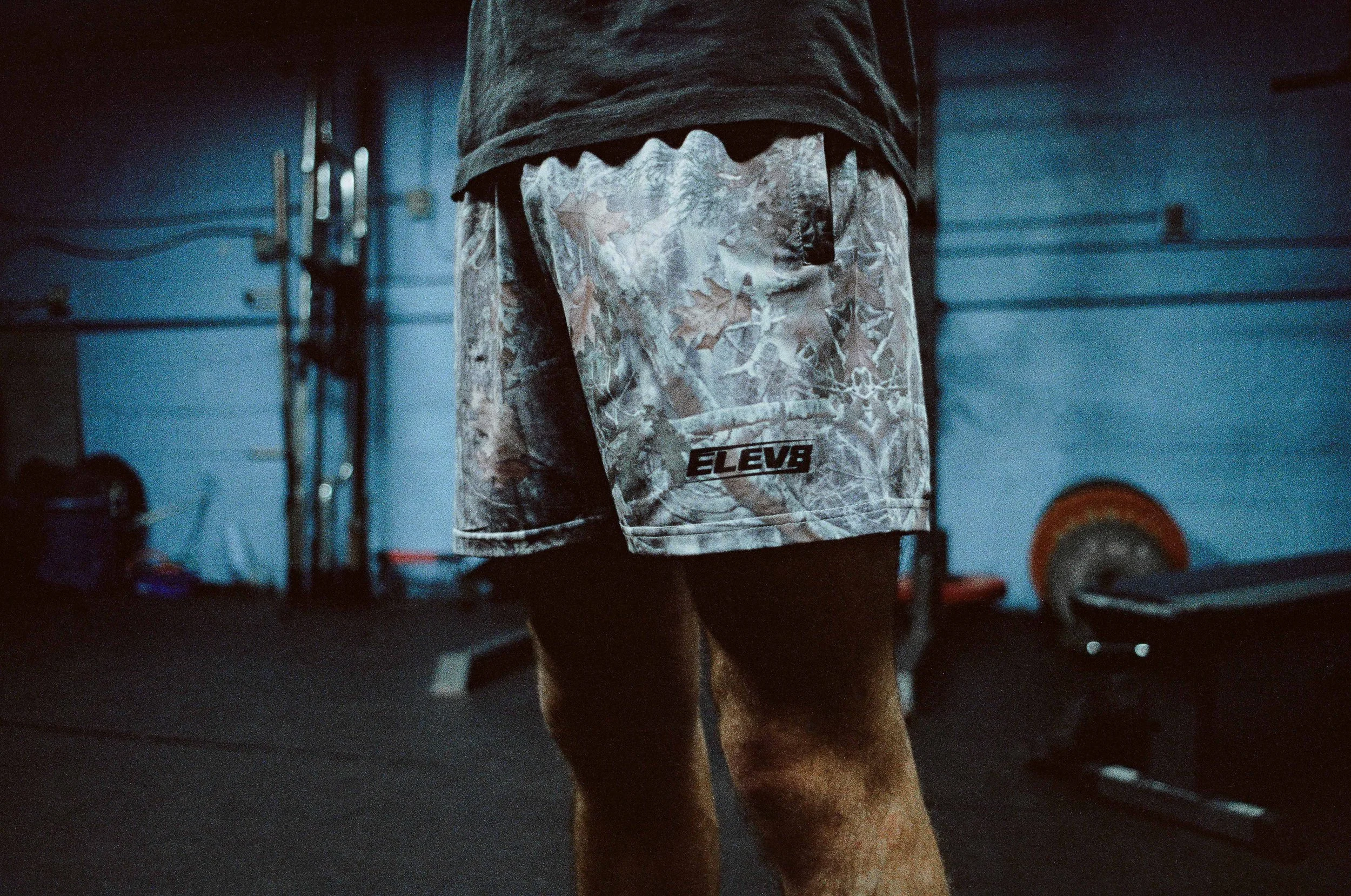 A person wearing camouflage shorts and a gray hoodie standing in a gym with weights and equipment in the background.