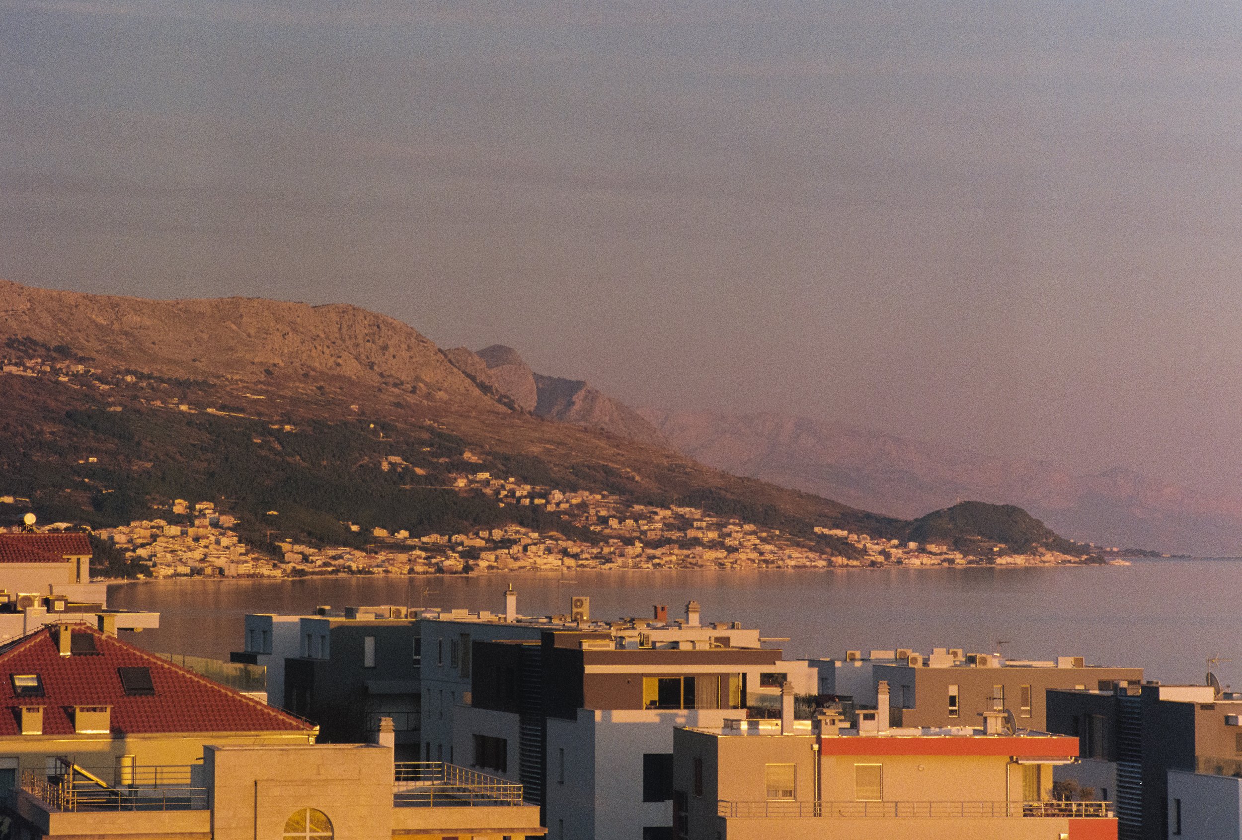 Sunset view of modern buildings overlooking a calm body of water with a hillside dotted with houses in the background.
