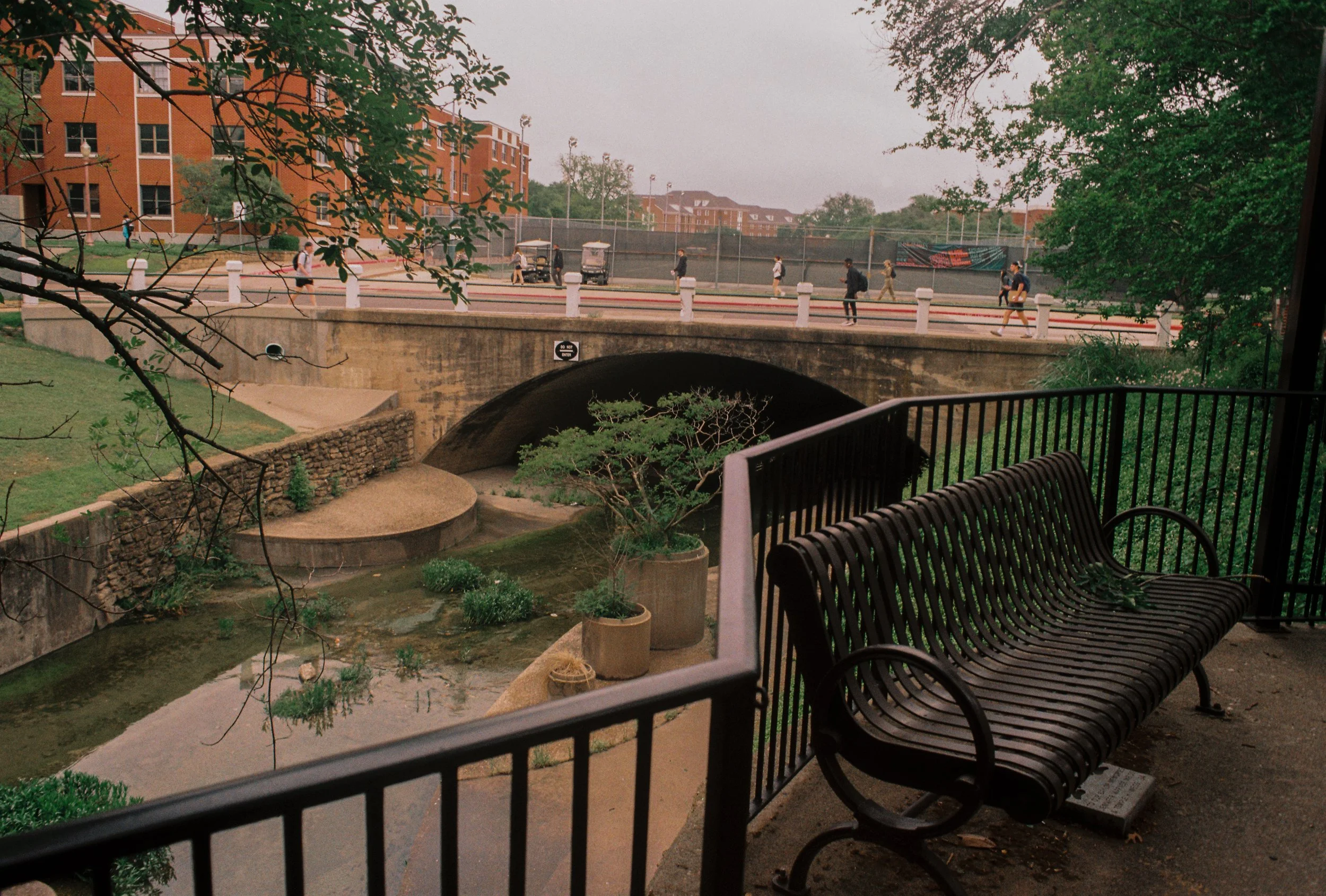 A park bench in the foreground overlooking a small pond and a bridge. People walk along the overpass in the background, with residential buildings and trees around.