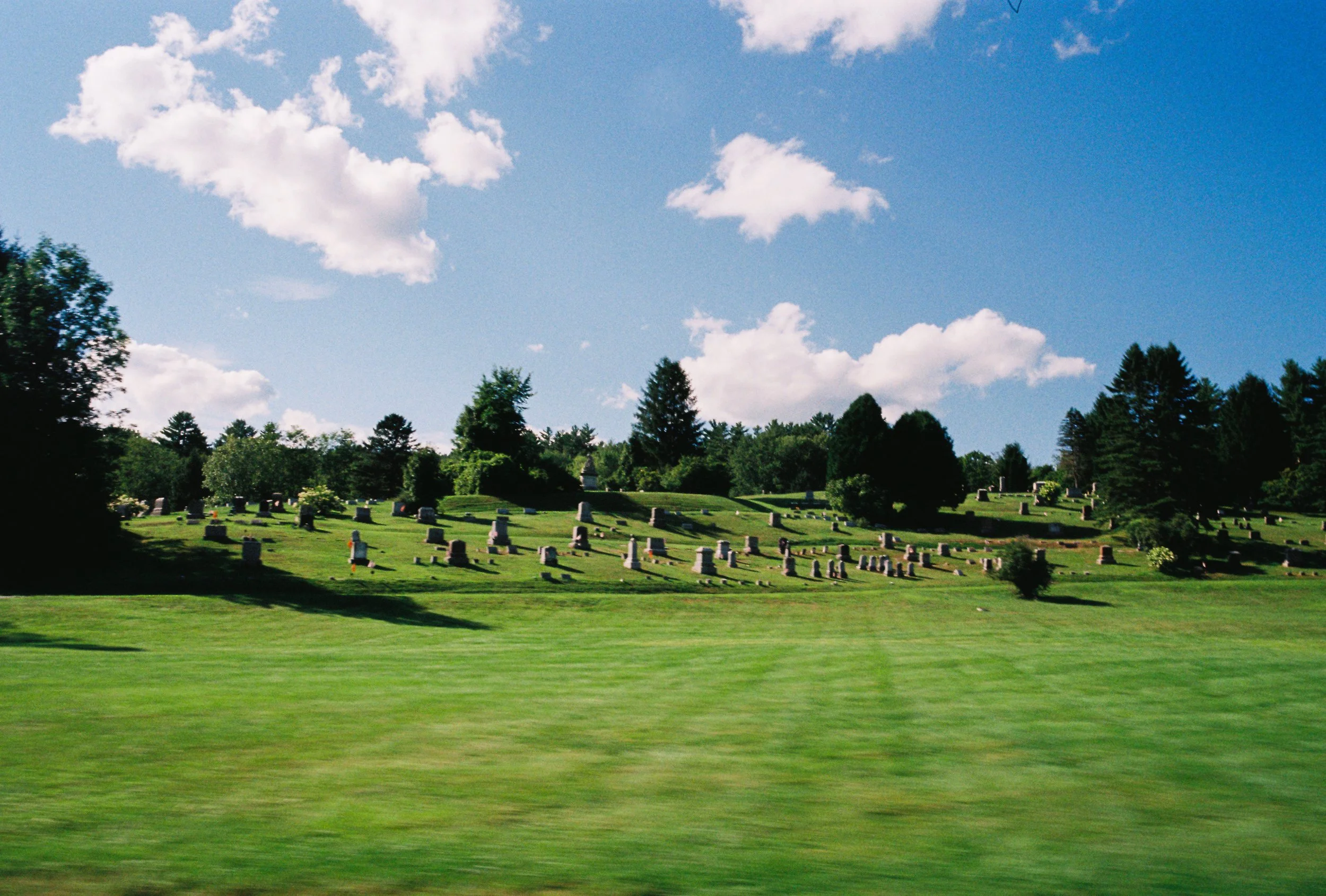 A cemetery on a hill, with numerous tombstones surrounded by green grass and trees, under a blue sky with scattered clouds.