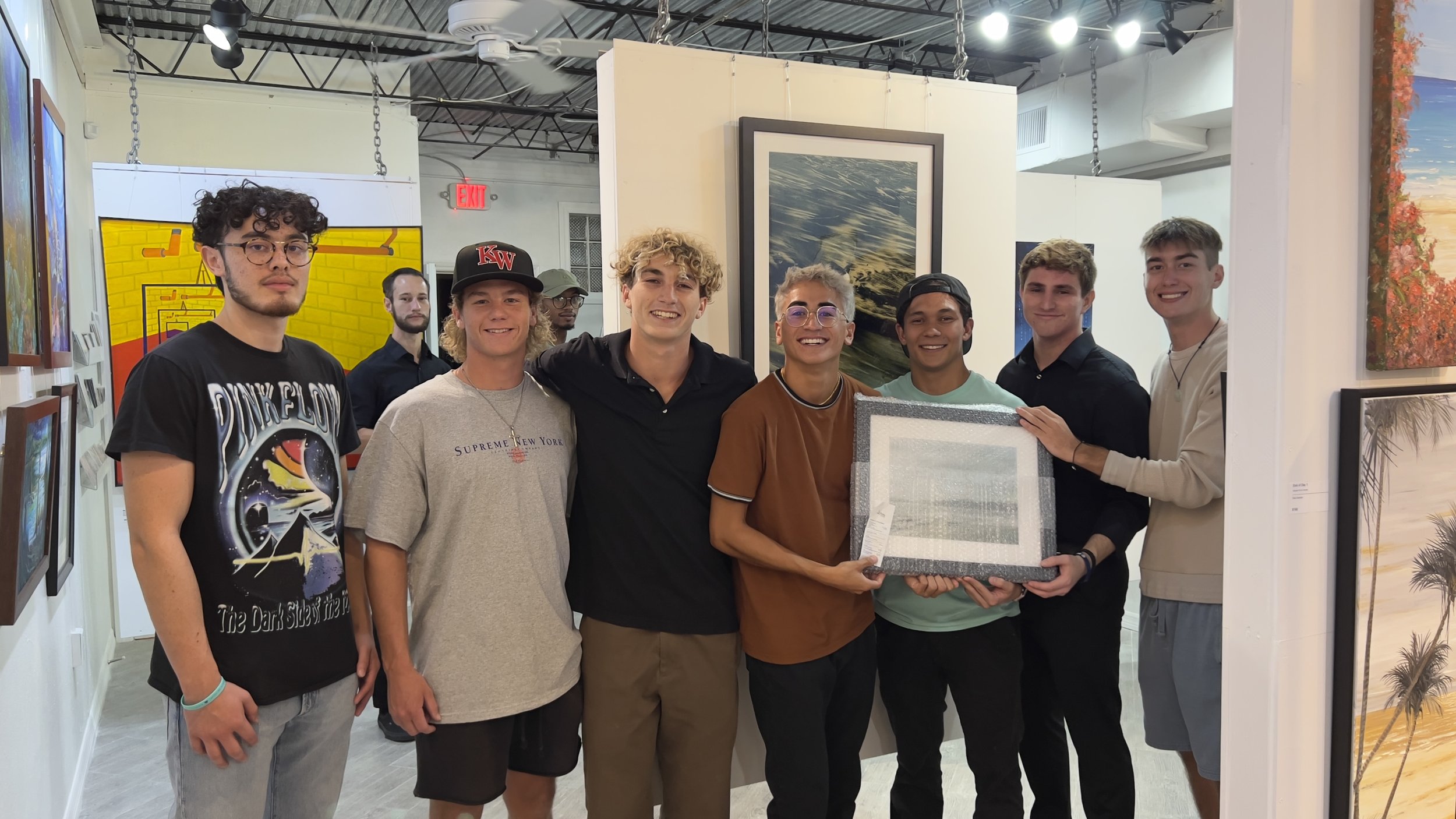 Group of eight young men standing in an art gallery, smiling, with some holding a framed artwork.