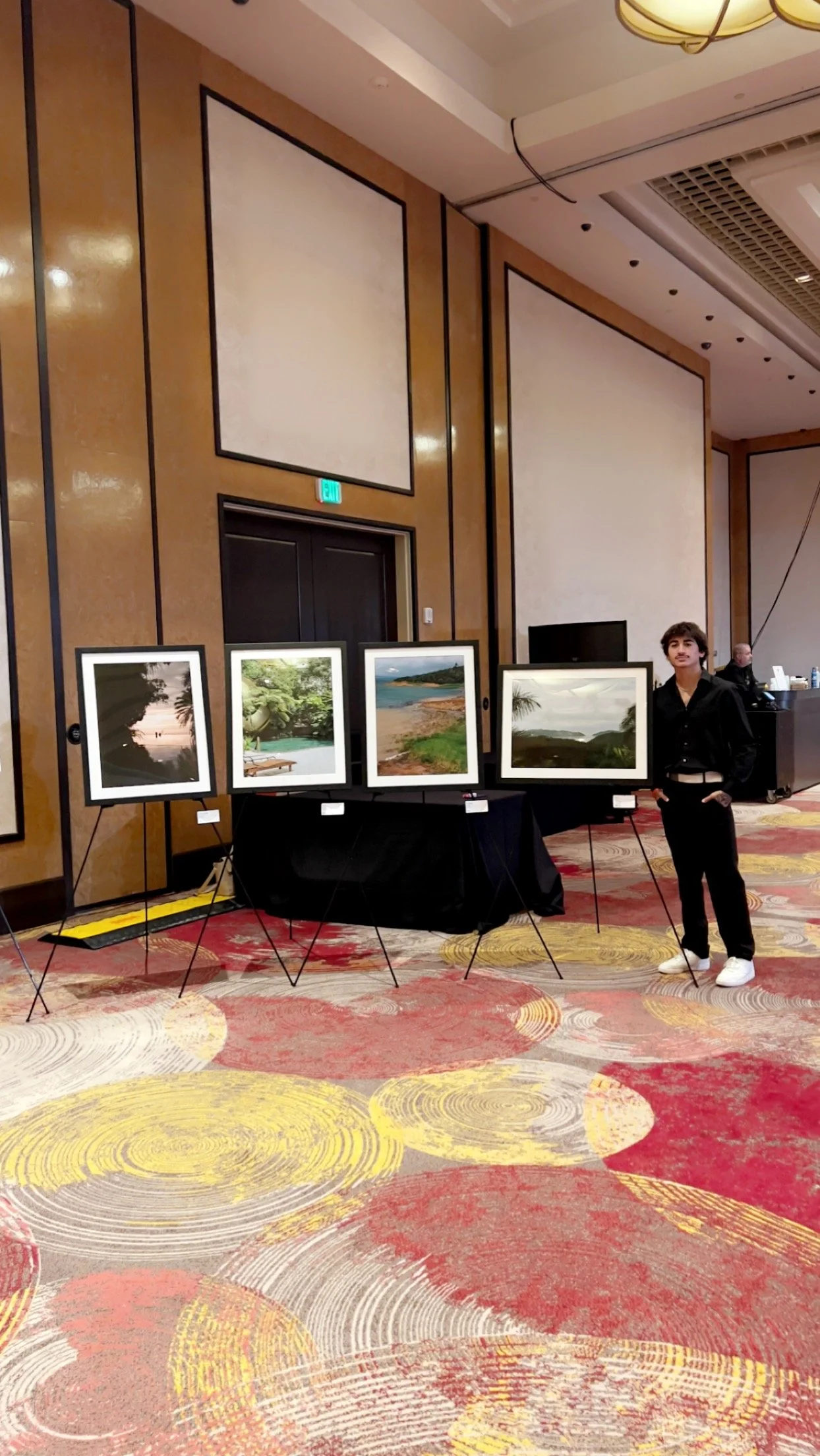 Gallery display with four framed landscape photographs on black stands and a person standing beside them in a large, indoor exhibition space with patterned carpet and beige walls.