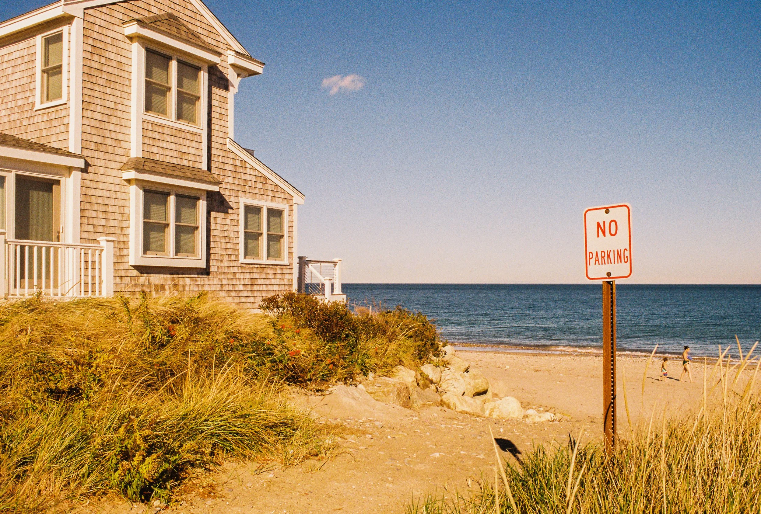 Beach house with wooden siding overlooking the ocean, with a 'No Parking' sign in the sand and a woman walking with children along the shoreline.