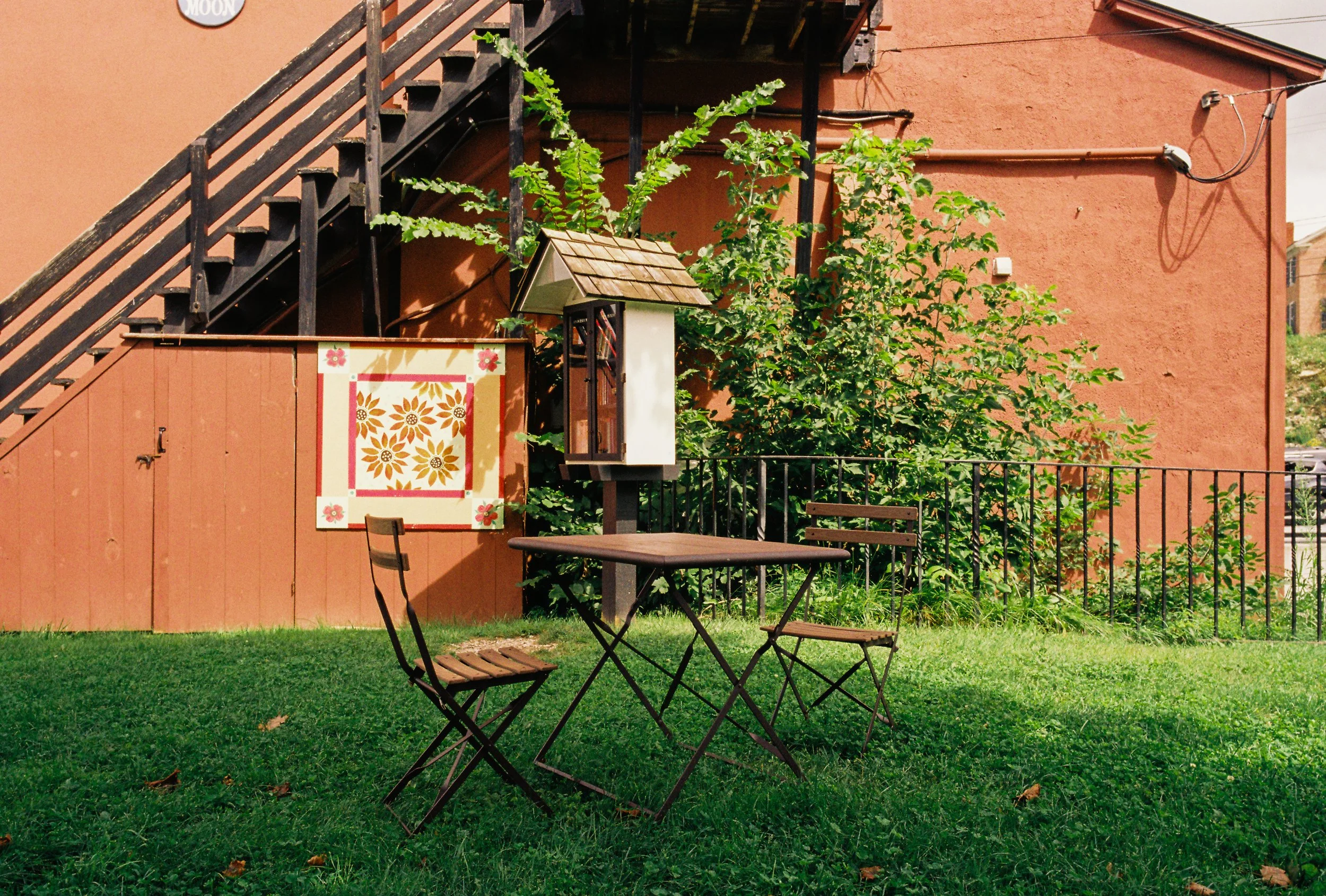 Outdoor patio with a table and three chairs on a grassy yard, against a rust-colored building wall with a staircase, satellite dish, and greenery.