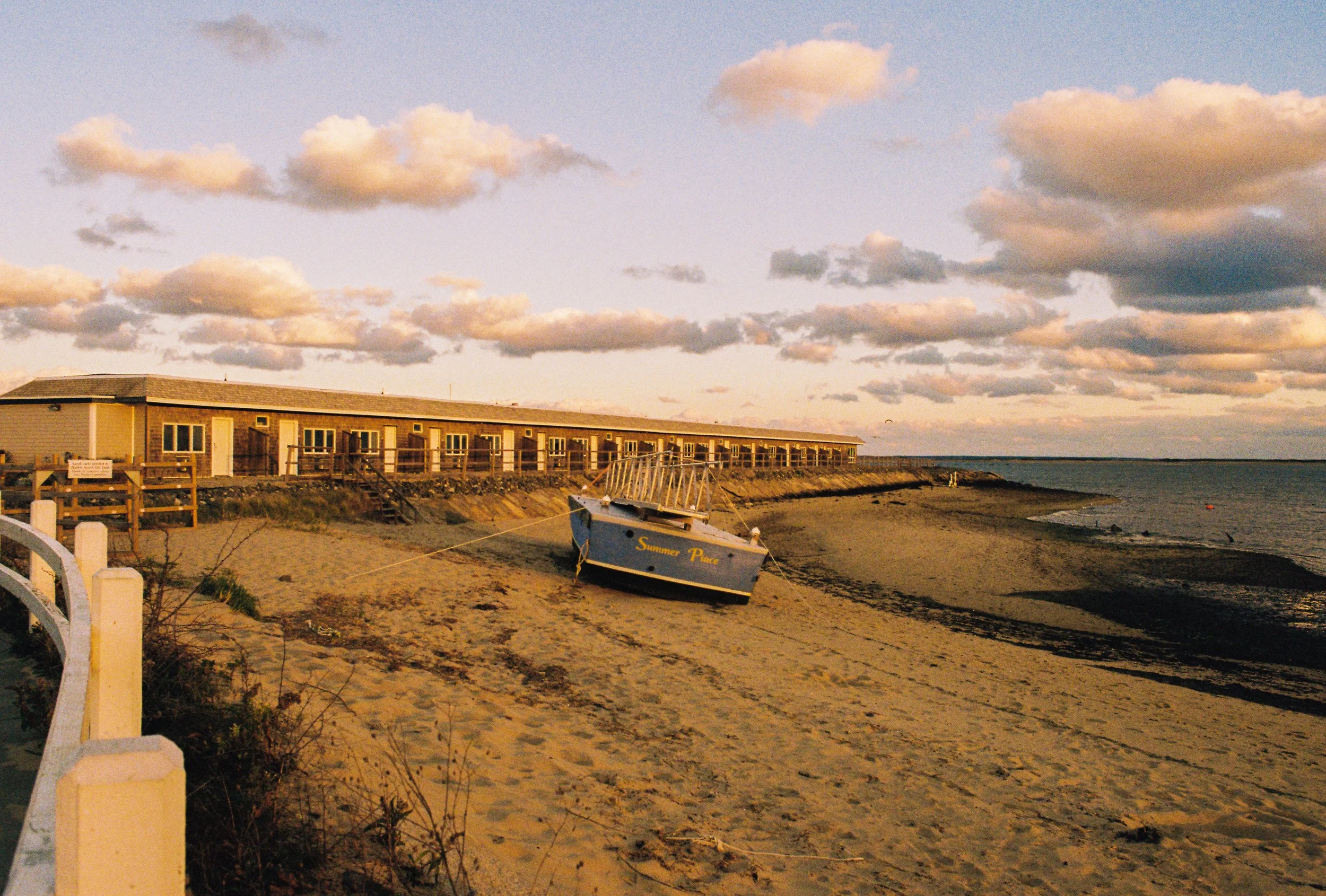A sandy beach with a small blue boat named 'Summer Place' resting on the shore, tied to a post. In the background, there are beach cabins or motel units, and a sky filled with scattered clouds during sunset or sunrise.