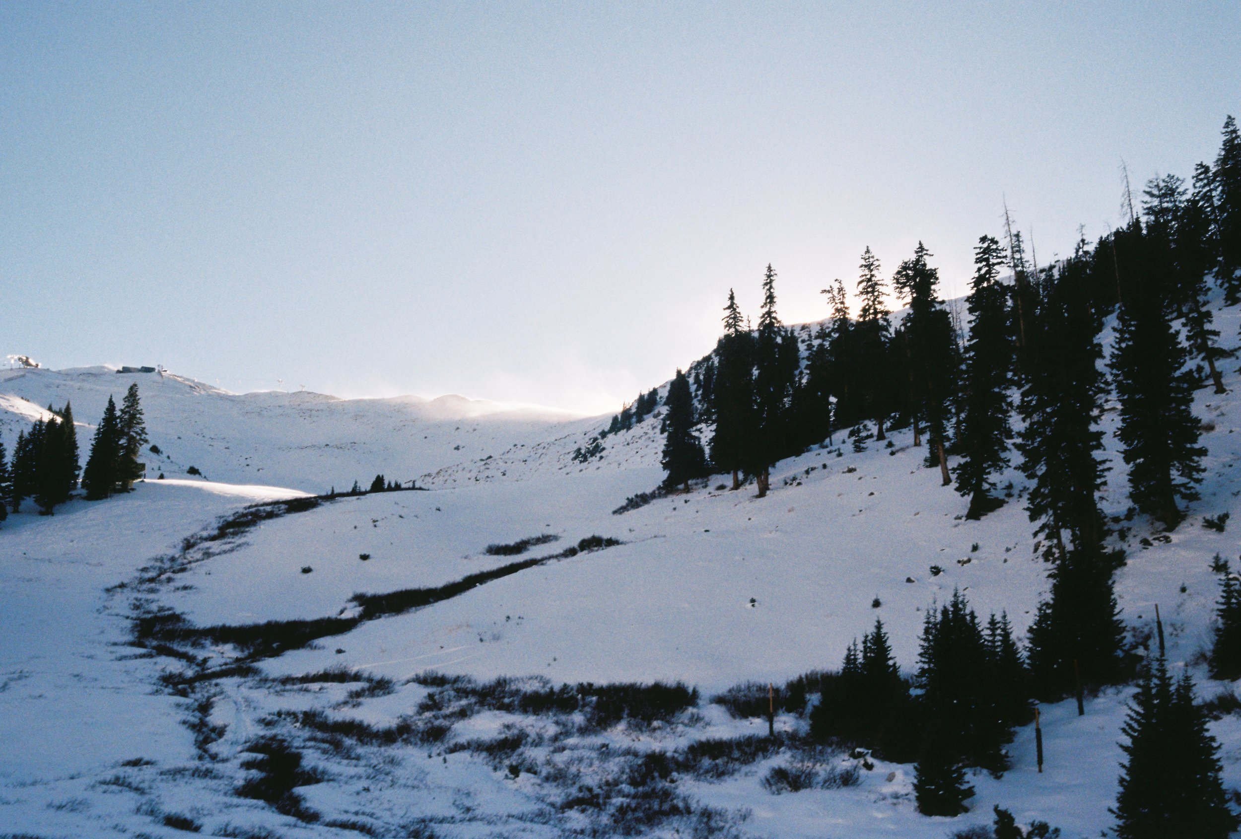 Snow-covered mountain landscape with scattered pine trees and a narrow trail winding uphill, under a partly cloudy sky.