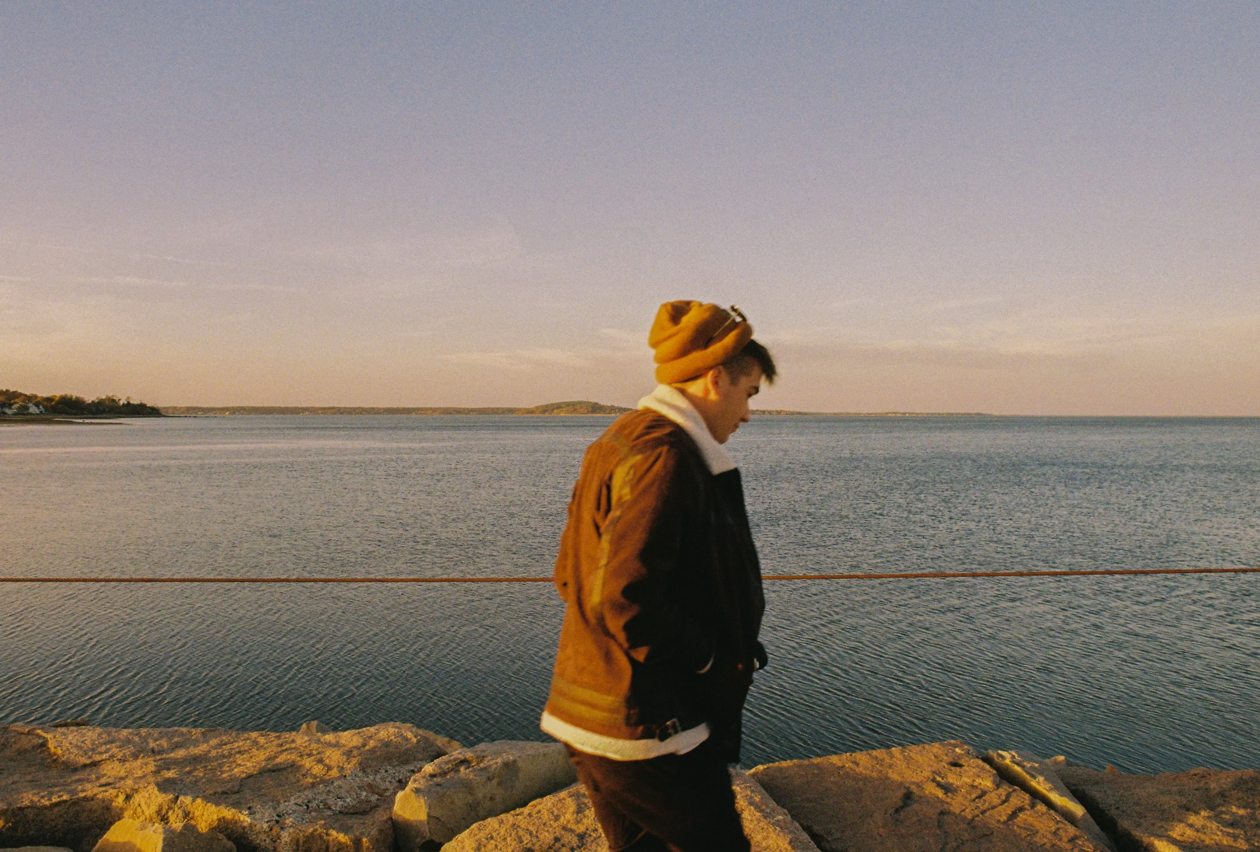 A young man wearing a brown beanie, black jacket, and white hoodie walking along a rocky shoreline near a body of water during sunset.