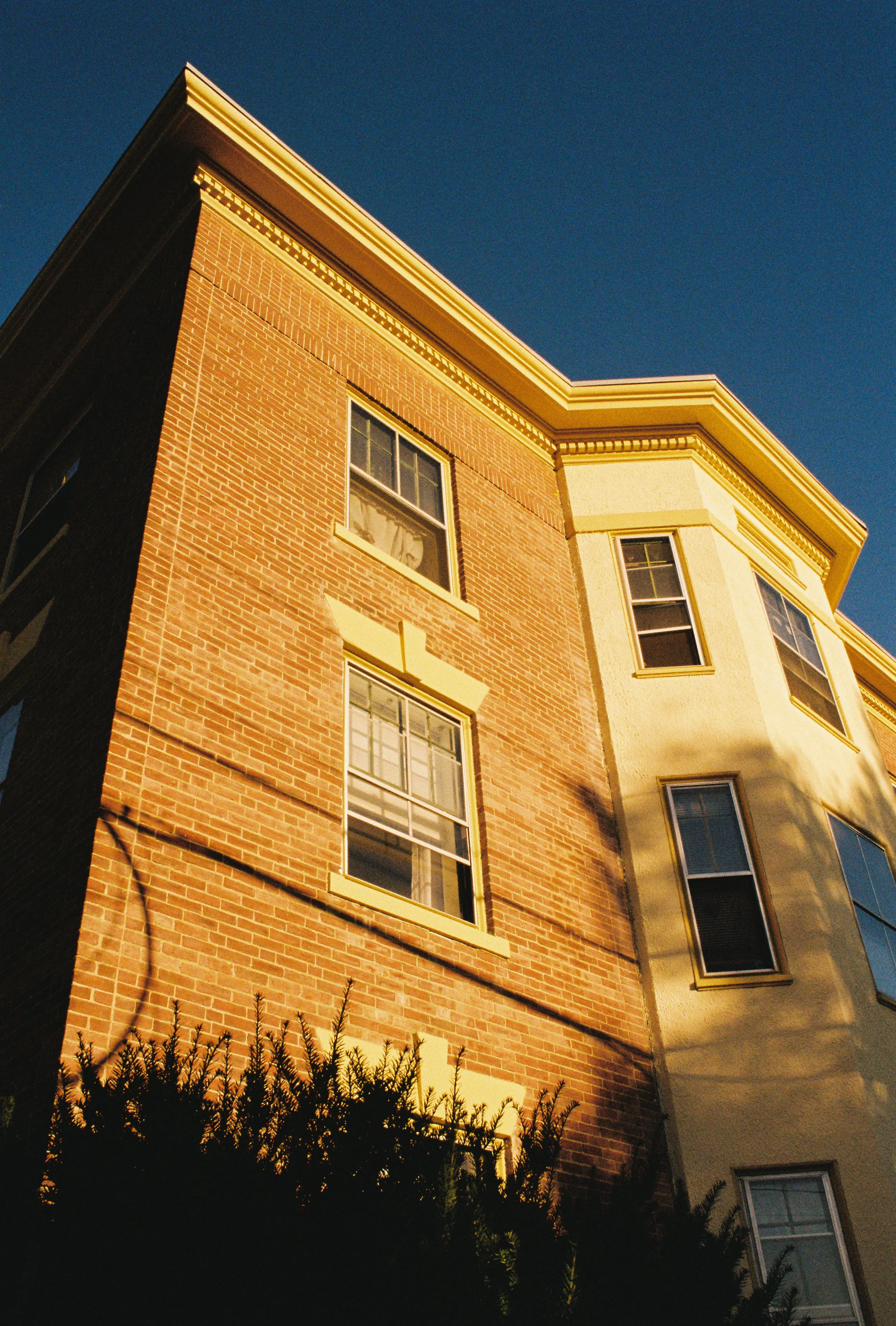 Low-angle view of a multi-story brick building with white trim, set against a clear blue sky in the late afternoon or early evening.