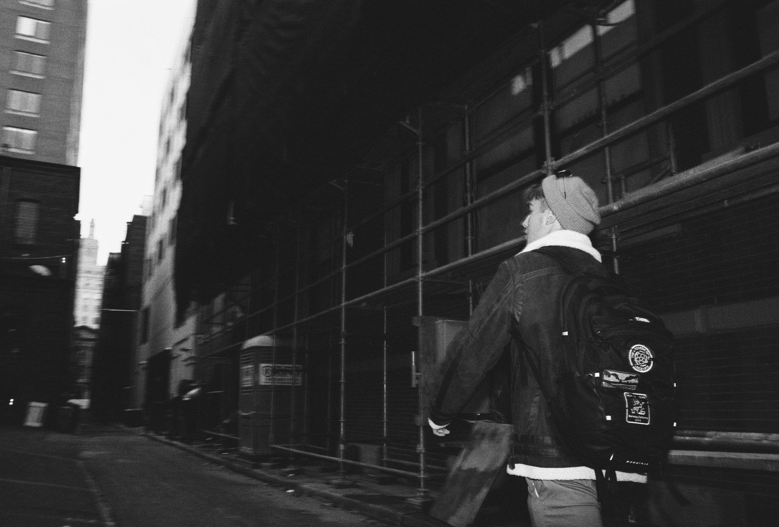 A young man wearing a beanie, denim jacket, and backpack walking down a city street at night, with tall buildings and construction scaffolding in the background.
