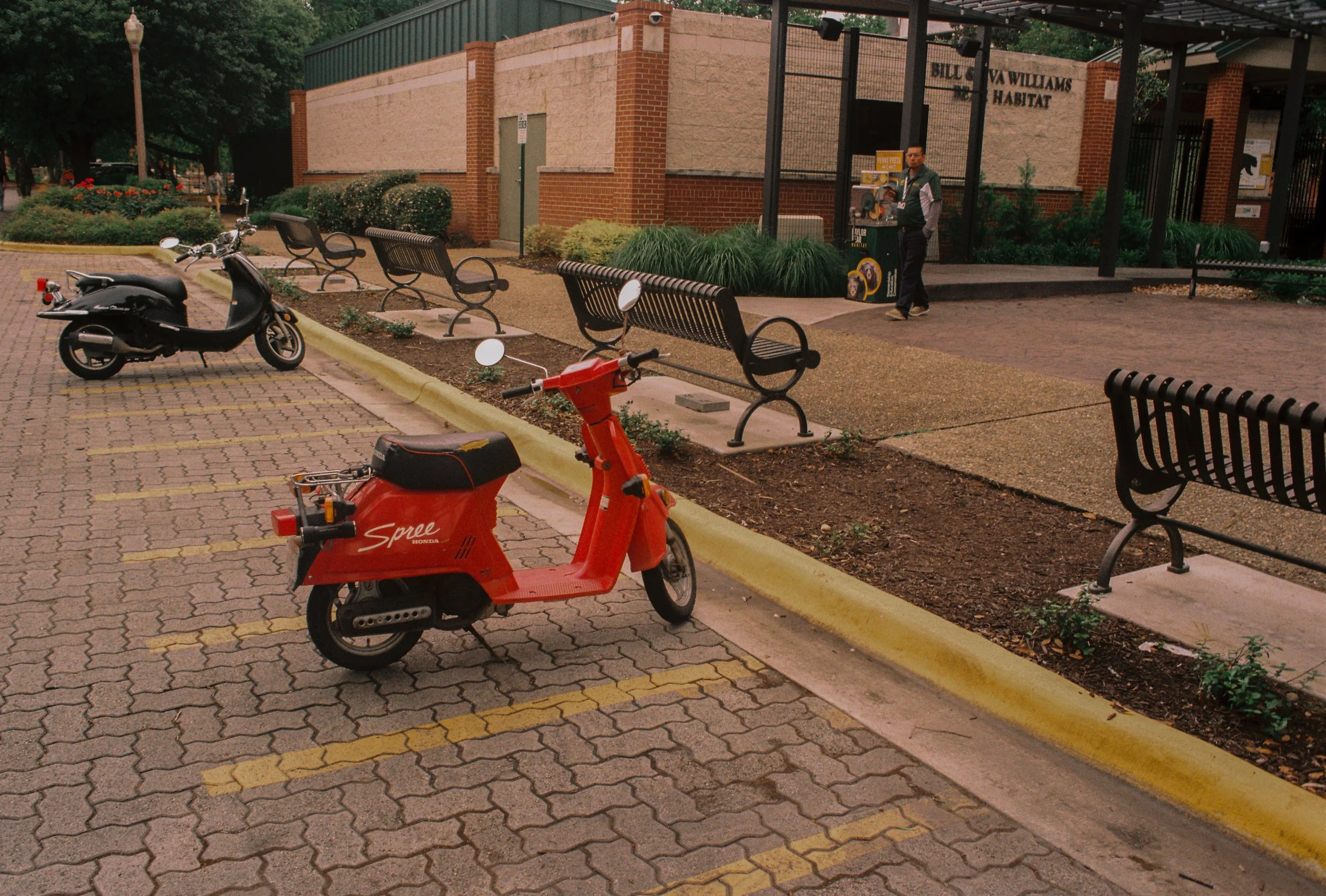 A parking lot outside an animal habitat exhibit with two scooters parked, benches, and a brick building with a sign that reads "Bill Oliver Williams Habitat". There is a person near the entrance and some greenery.