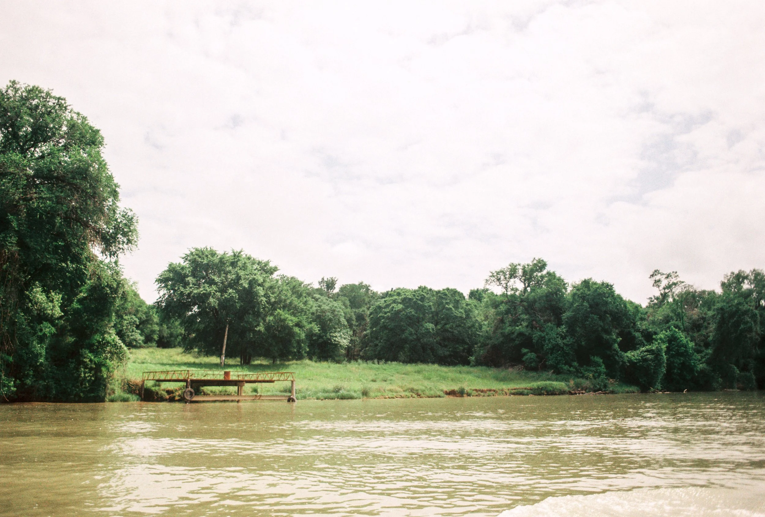 River with brown water and a green, tree-lined bank under a cloudy sky.