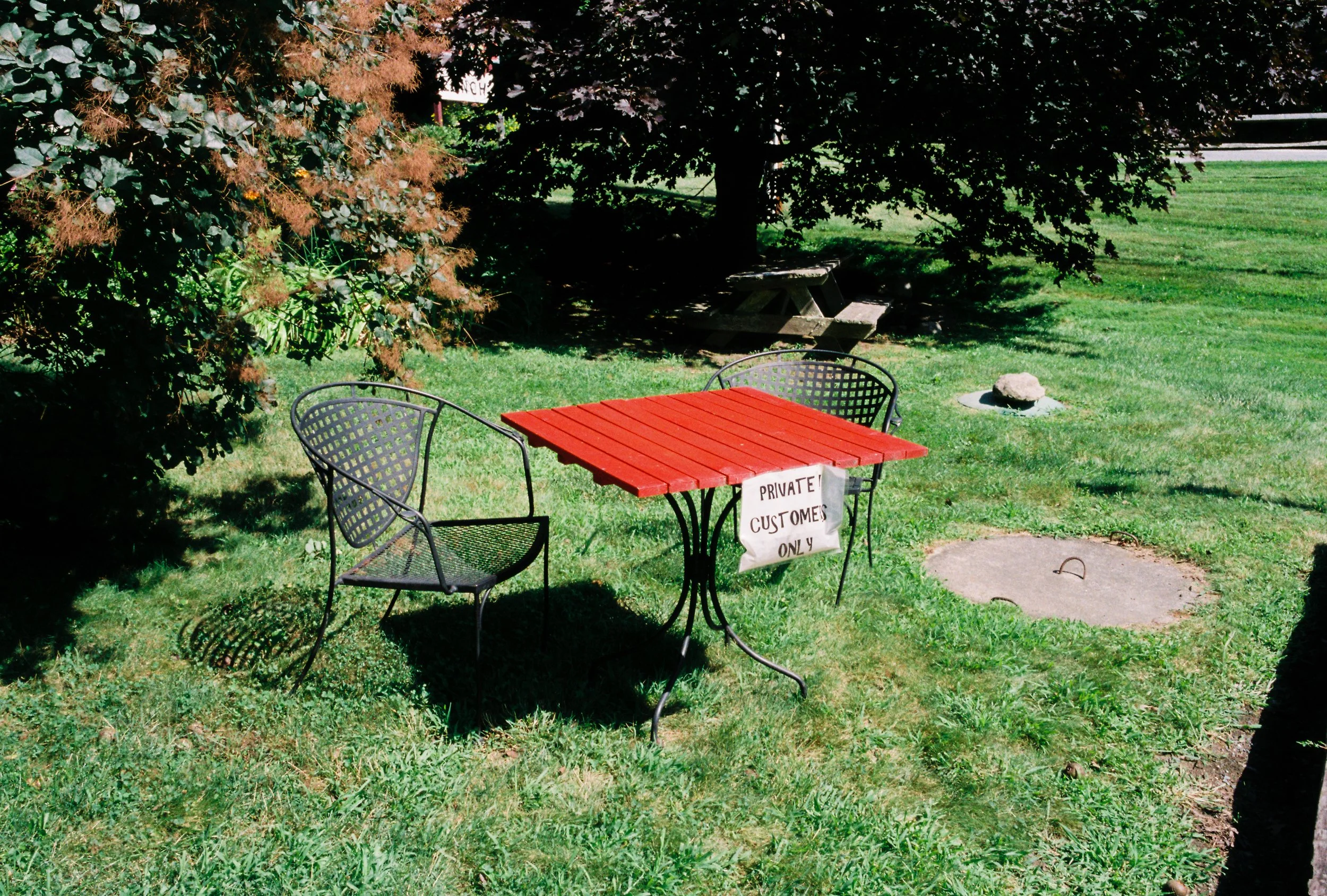 A small outdoor table with a red top and a handwritten sign that reads 'Private Customers Only' in black marker hanging from it, surrounded by three black metal chairs, in a green grassy area with trees and a picnic table in the background.