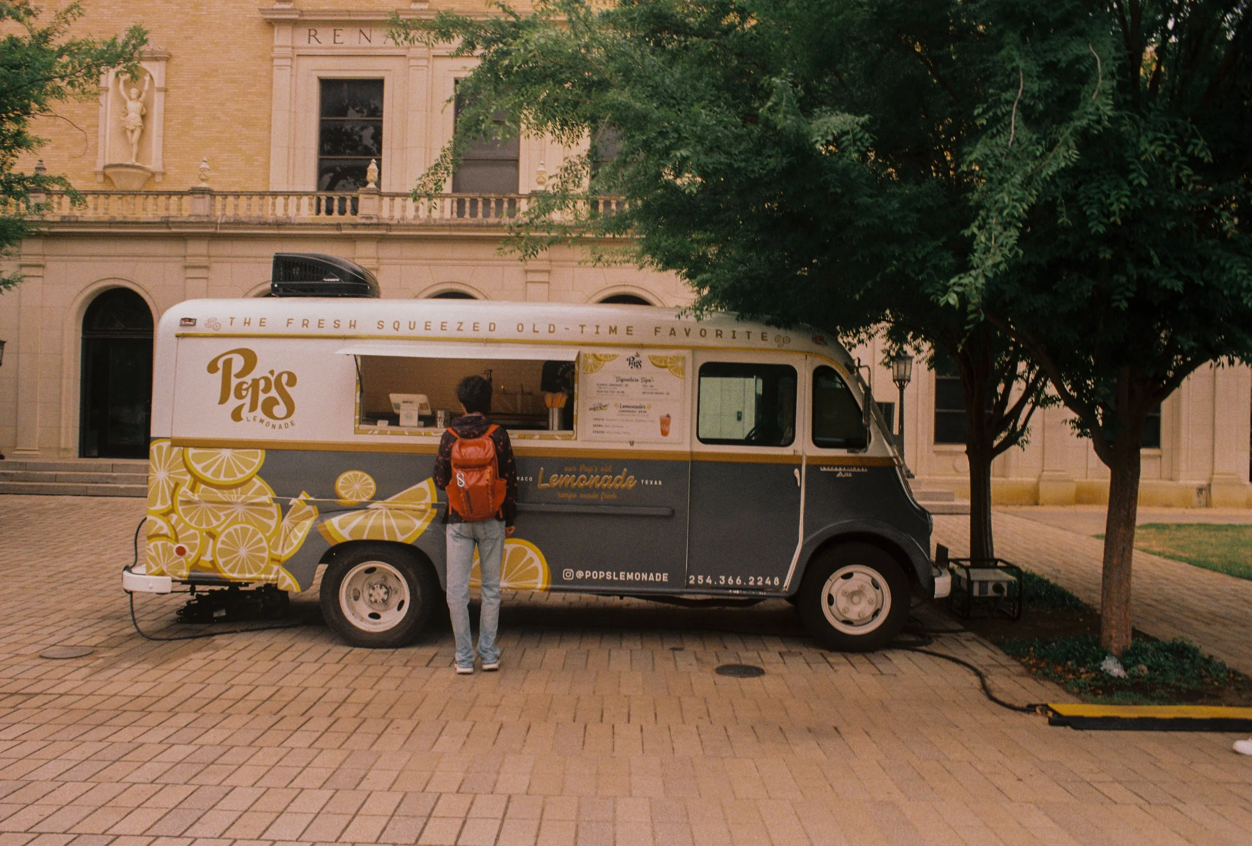 A person in jeans and a dark hoodie with a red backpack stands in front of a lemonade truck on a cobblestone street, with trees and a building in the background.