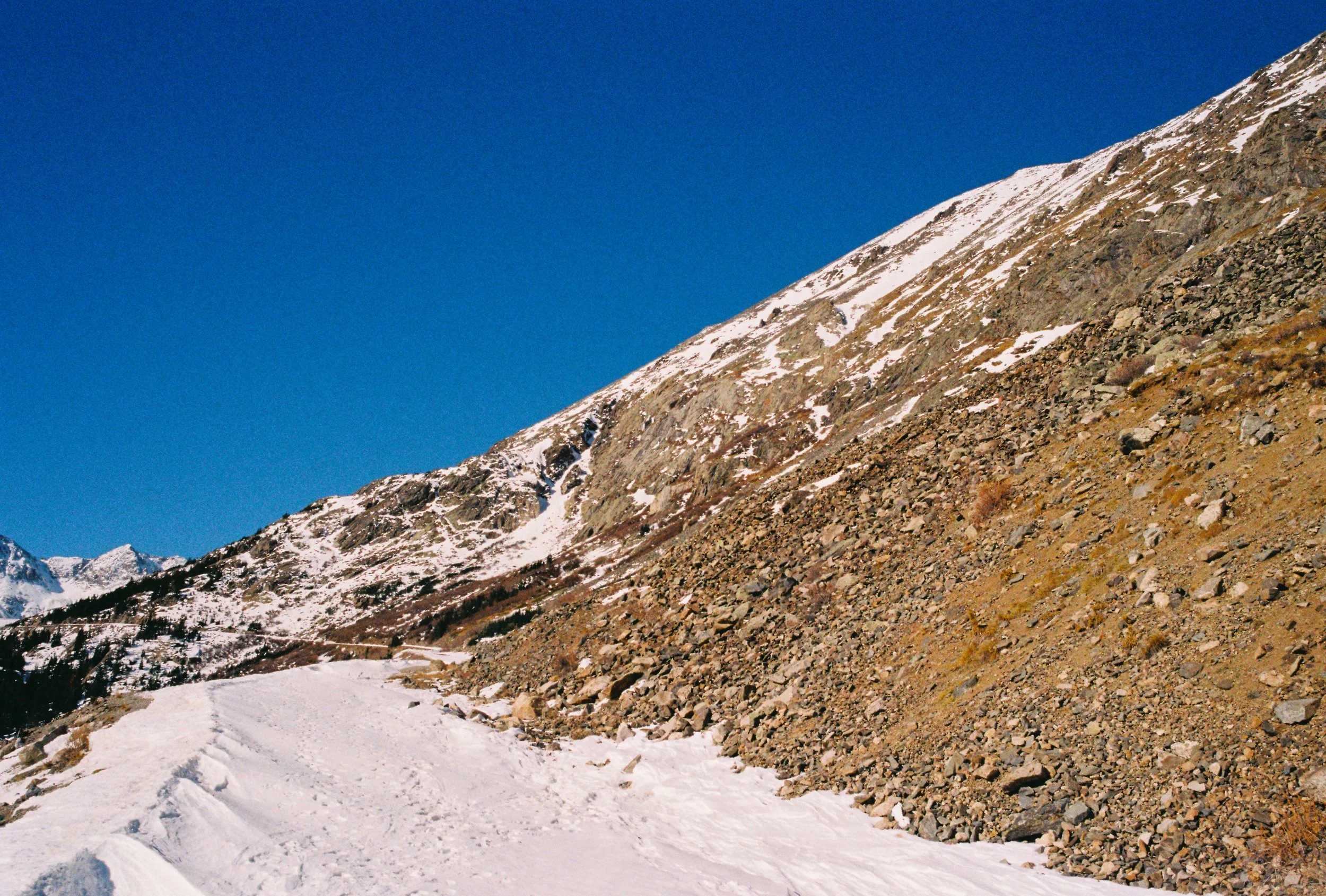 Snow-covered mountain trail with rocks on the side and snow patches on the slopes, under a clear blue sky.