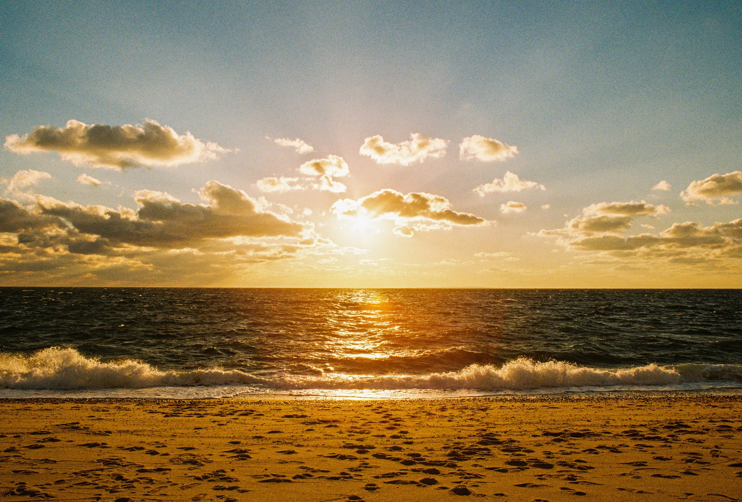 Sunset over the ocean with sandy beach and scattered clouds in the sky.