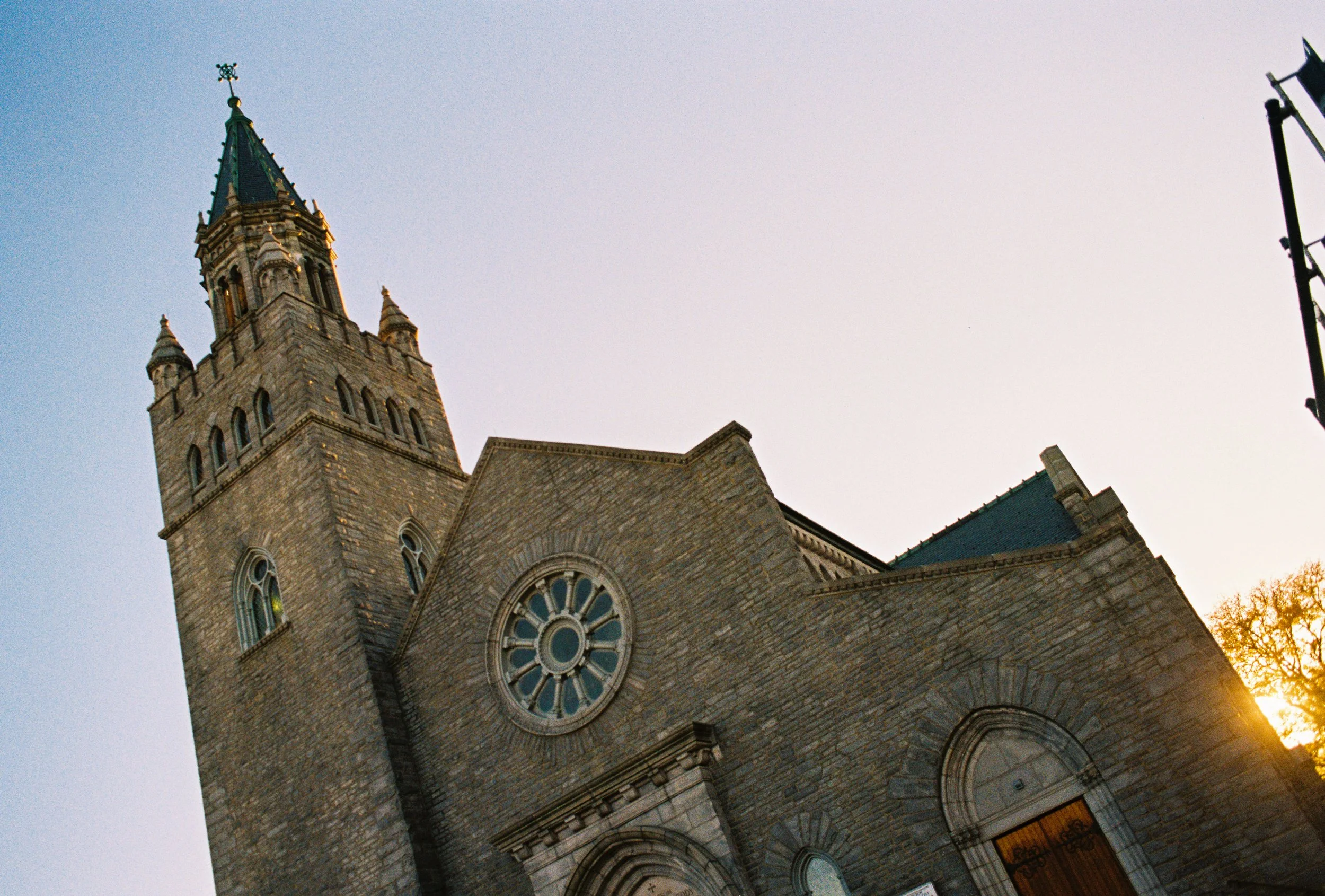 A historic stone church with a tall steeple and round stained glass window, captured at sunset with sunlight peeking from the right side.