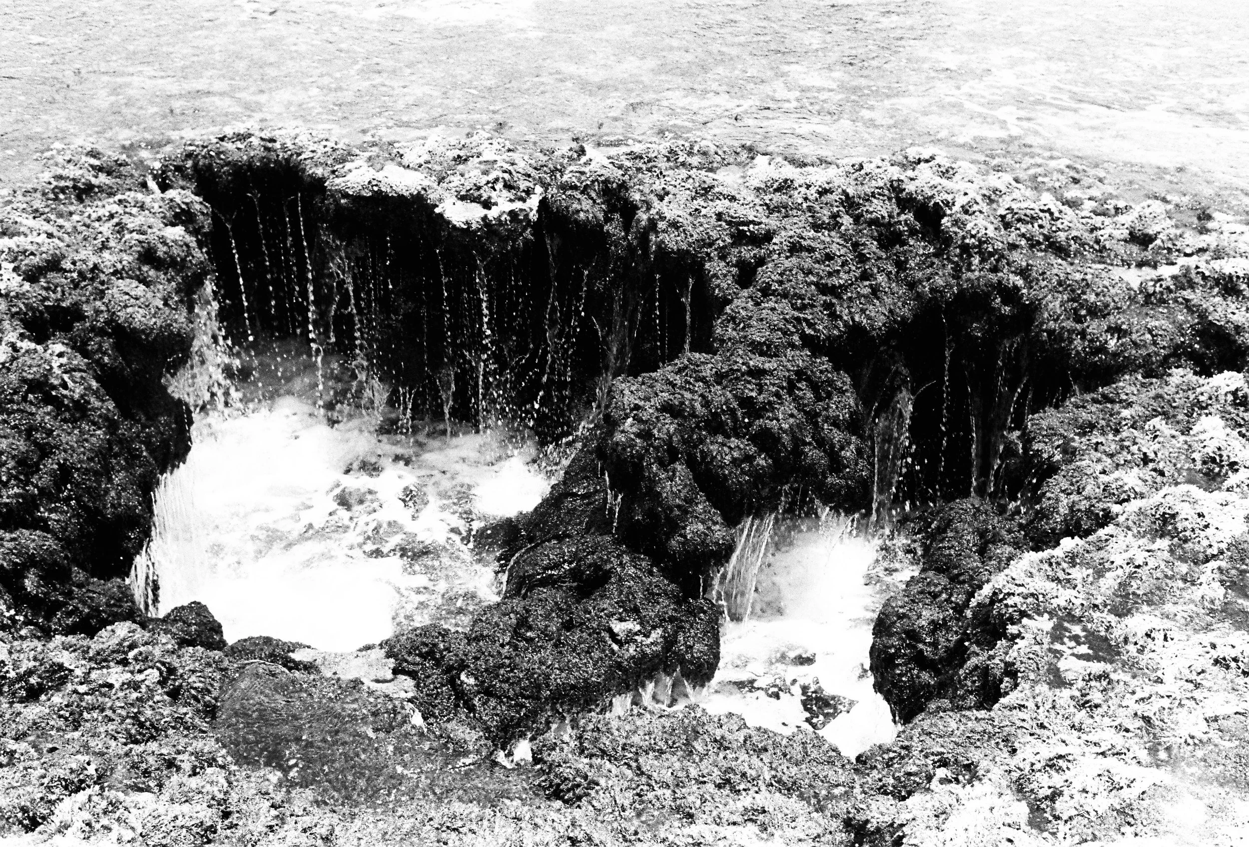 Black and white photo of a small tidal pool or blowhole on a rocky shoreline with water cascading over rocks.