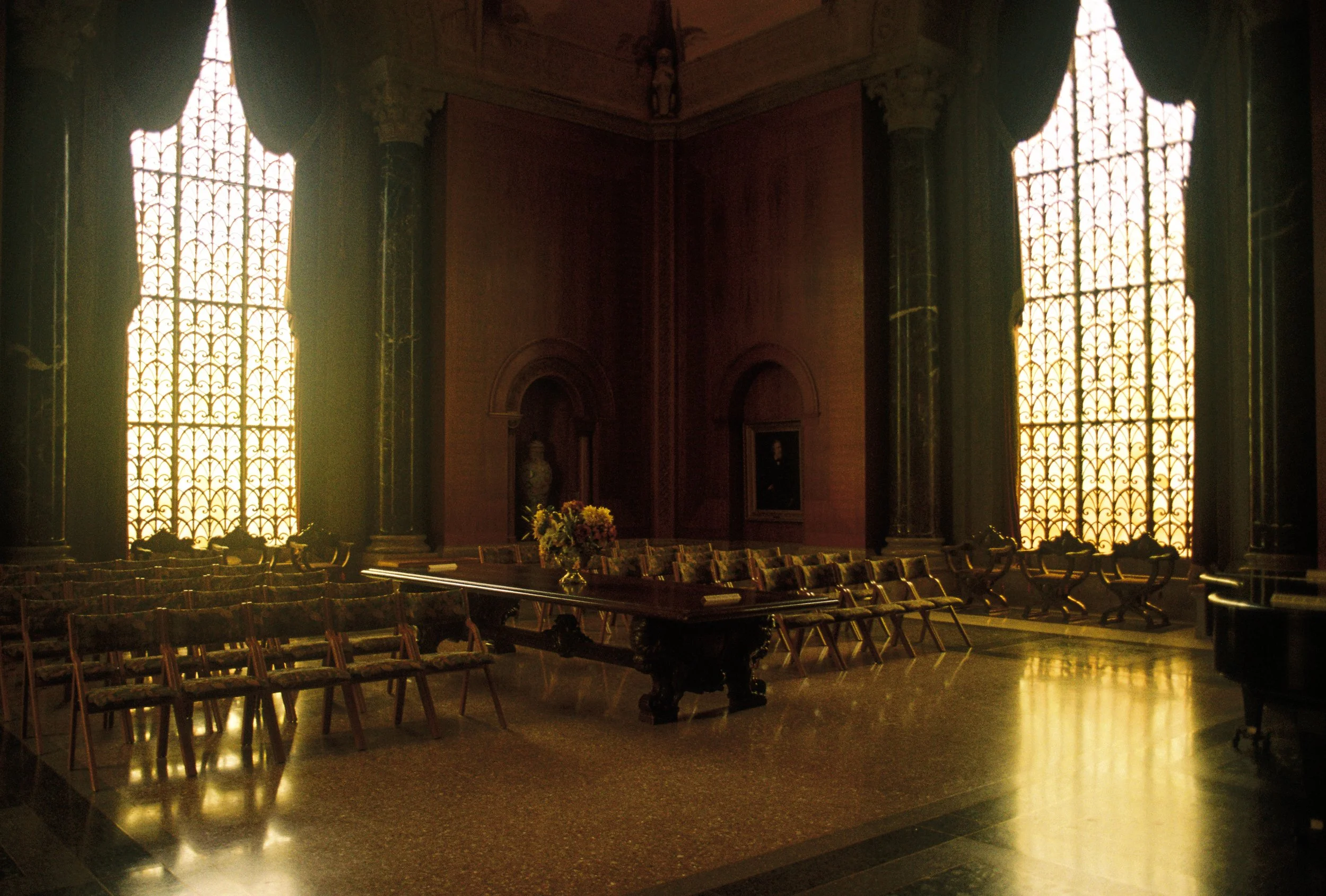 A large, historic room with tall, ornate windows allowing sunlight to glow inside. The room has dark columns, a polished floor, and a large wooden table with a floral arrangement. There are chairs around the table and along the walls, with some artwo