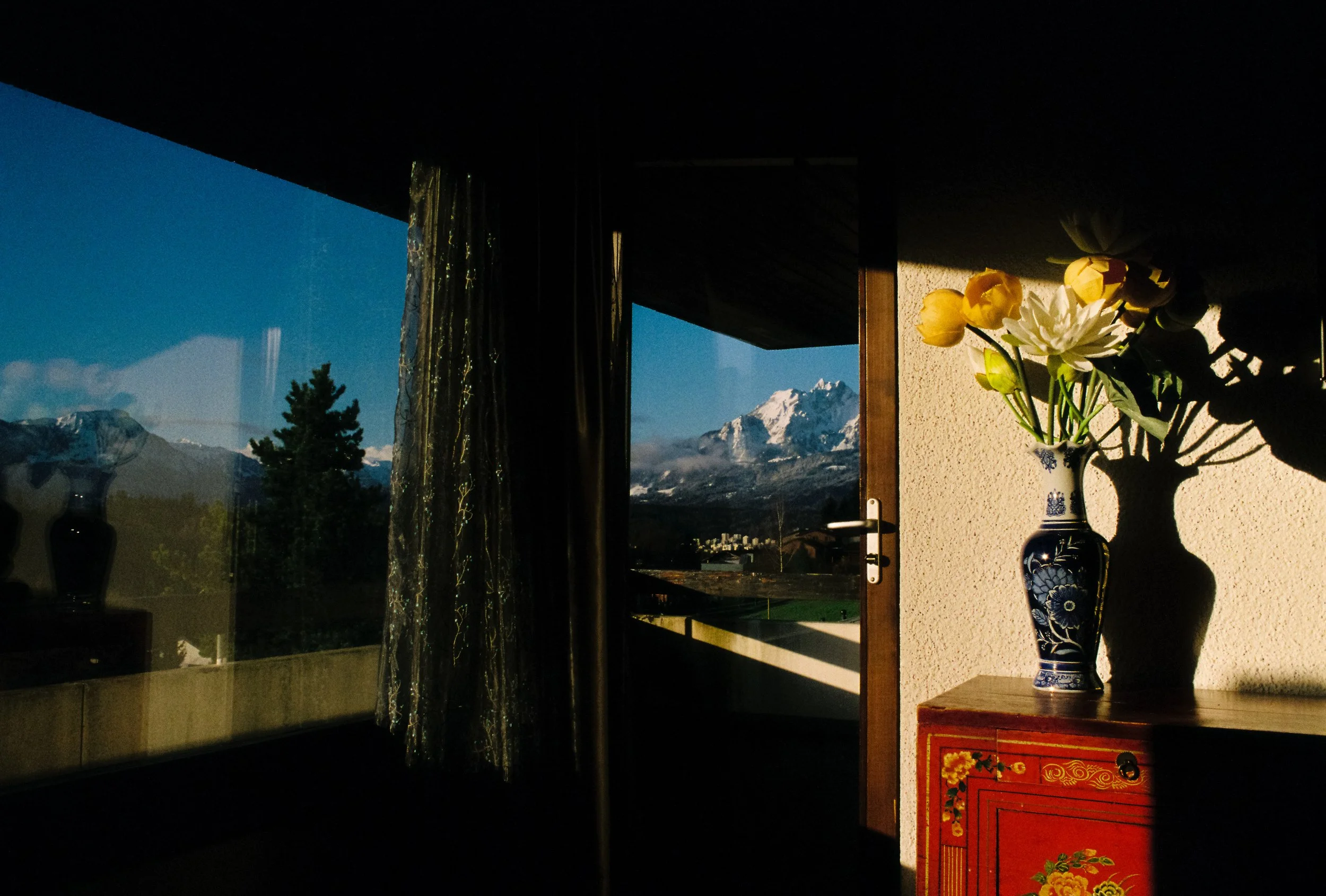 Sunlight illuminates a room with a blue and white vase of flowers on a red cabinet, casting shadows on the wall. Outside is a mountain landscape with snow-capped peaks under a clear blue sky, seen through an open door and window.