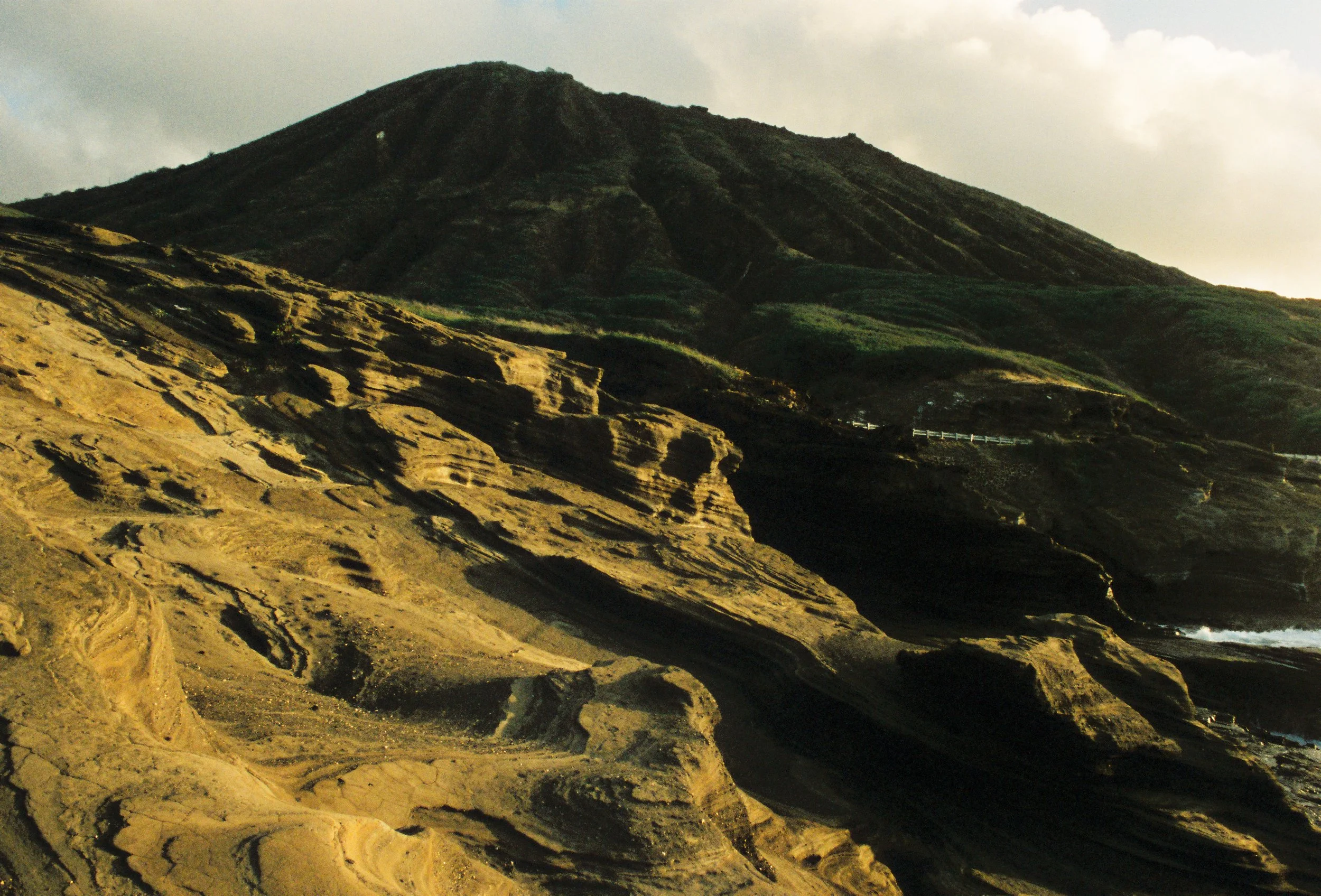 A rugged mountain landscape with layered rock formations and a dark volcanic mountain in the background under a cloudy sky.