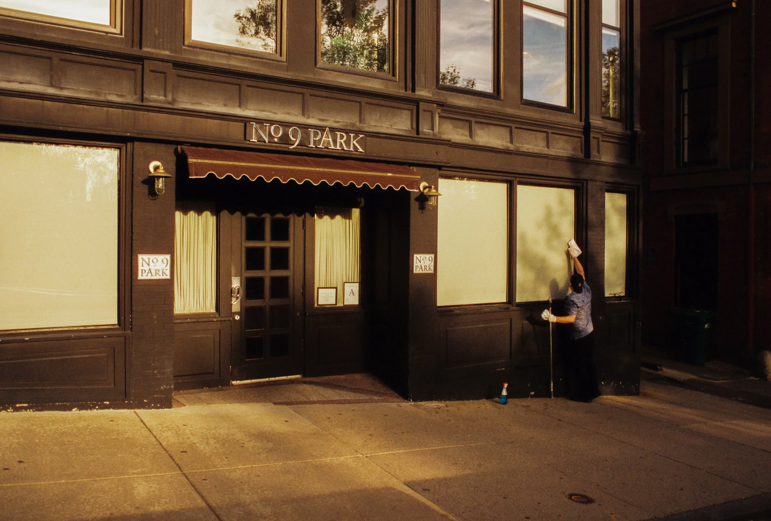 Man cleaning or painting exterior wall of a building labeled 'NO 9 PARK' with a squeegee or cloth, standing on sidewalk near a spray bottle.