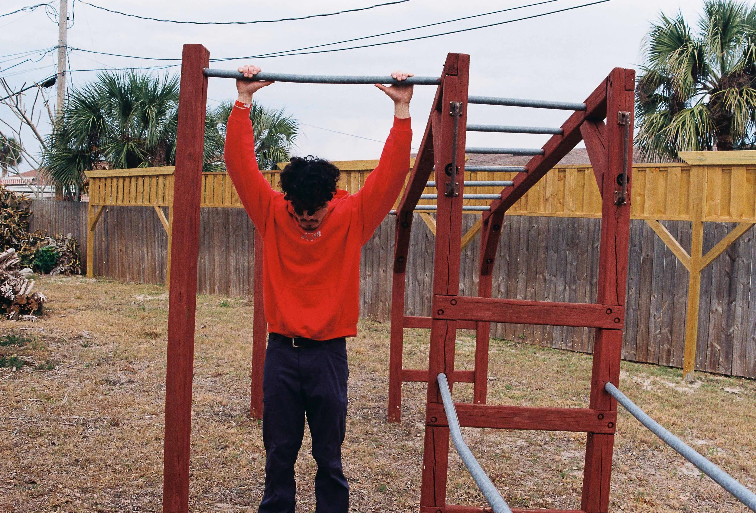 A person with curly hair wearing a red jacket and black pants is hanging upside down from a horizontal bar on outdoor playground equipment, with a wooden fence and palm trees in the background.