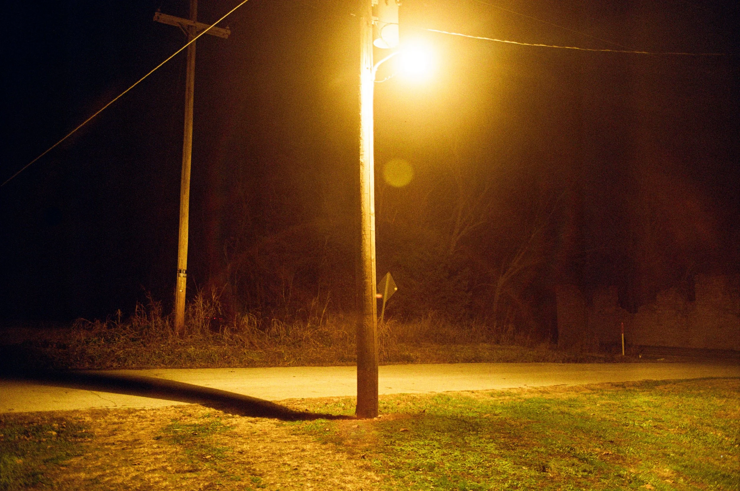 A wooden utility pole with a streetlamp on top illuminates a dark street at night, with grass and trees in the background.