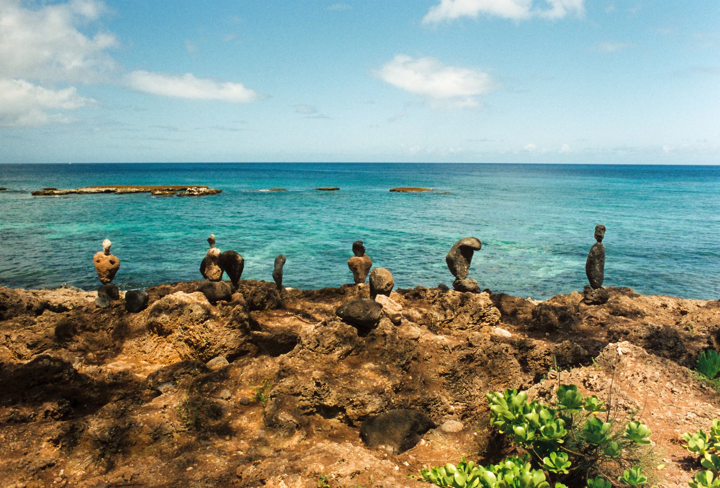 Stacked rocks along a rocky shoreline with blue ocean water and a cloudy sky in the background.